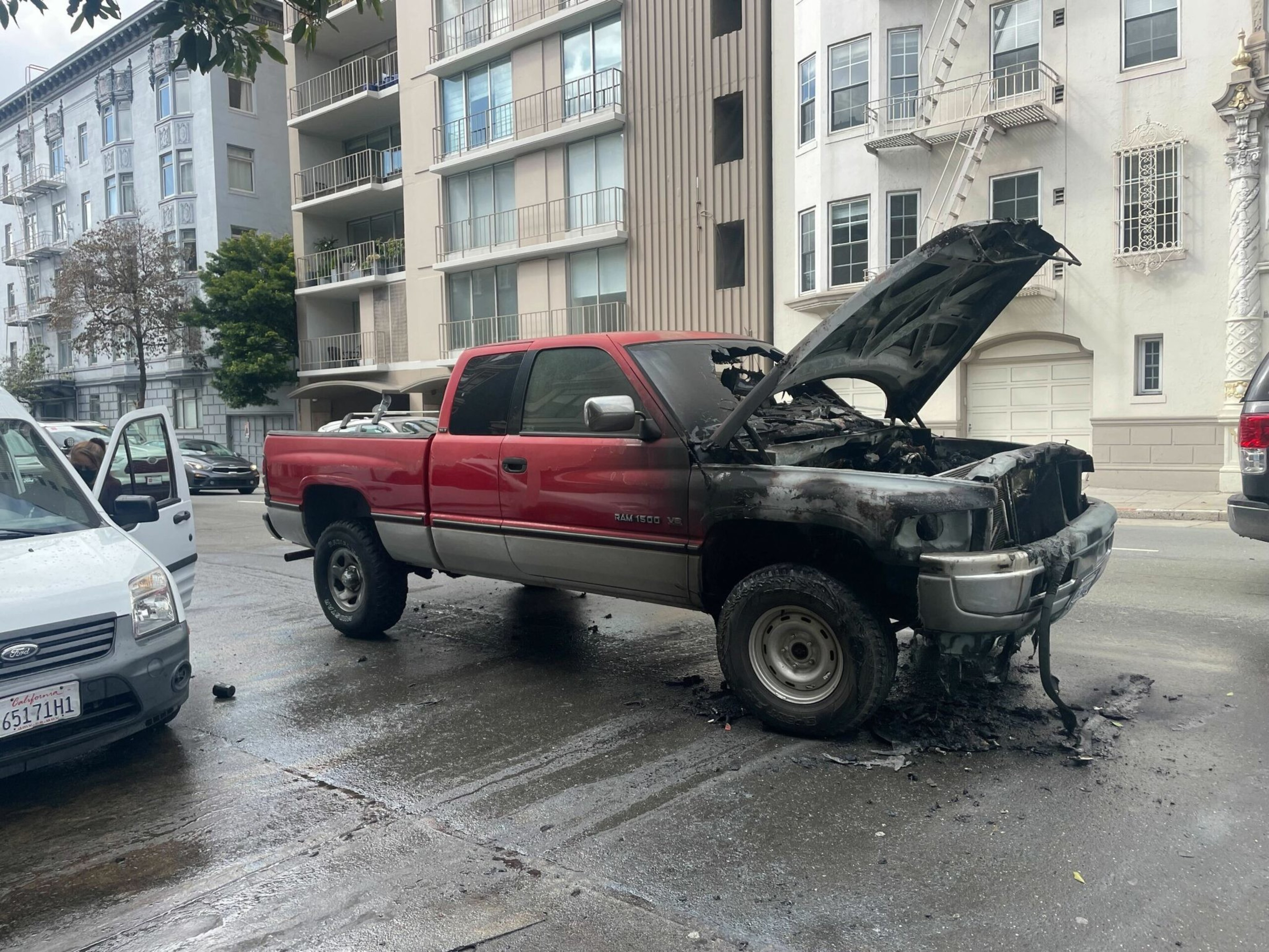 A red and gray pickup truck with severe fire damage to the front and an open hood is parked on a wet city street next to a white vehicle with an open door.