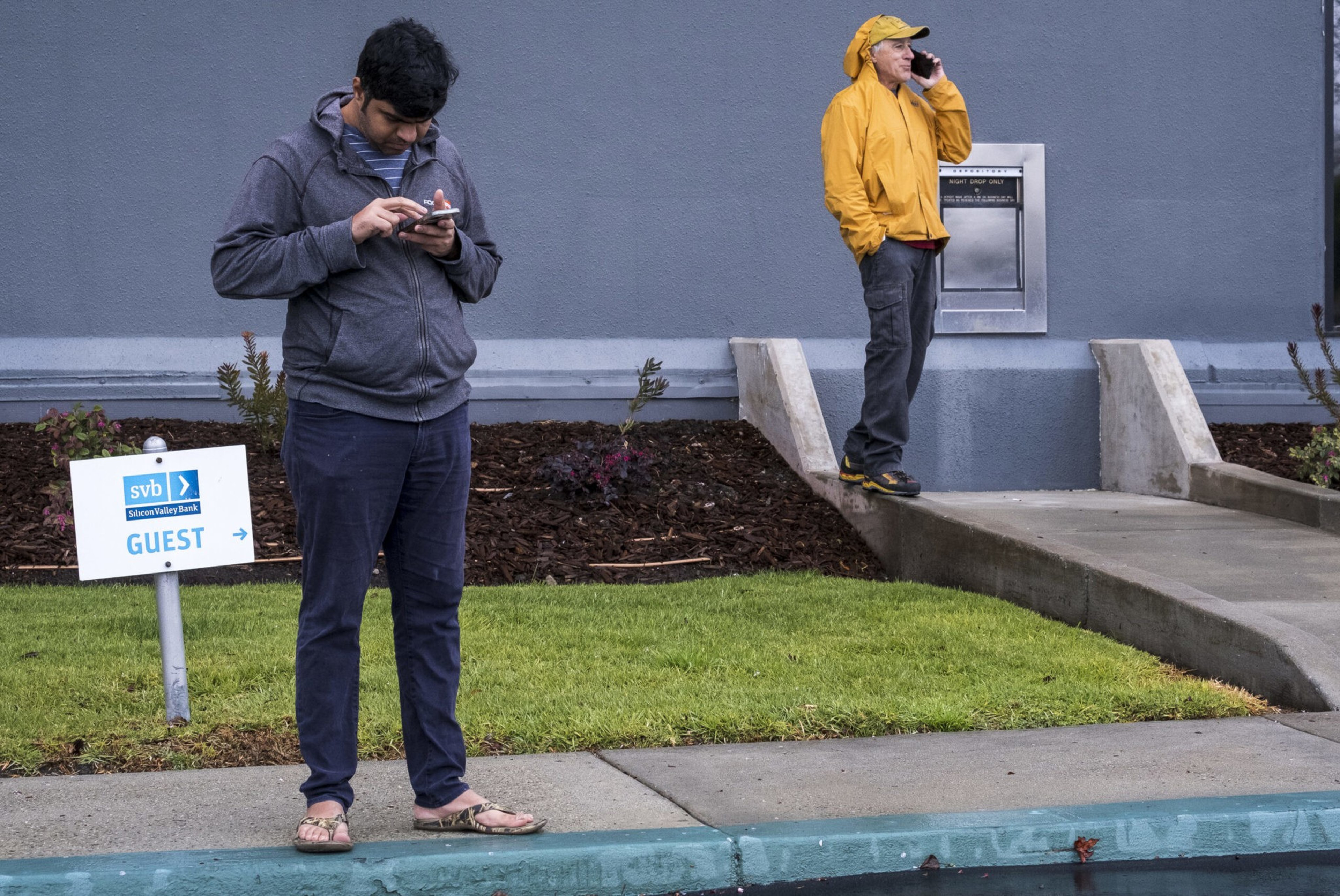 A man in a gray hoodie and sandals looks at his phone near a Silicon Valley Bank guest sign while another man in a yellow jacket talks on a phone.