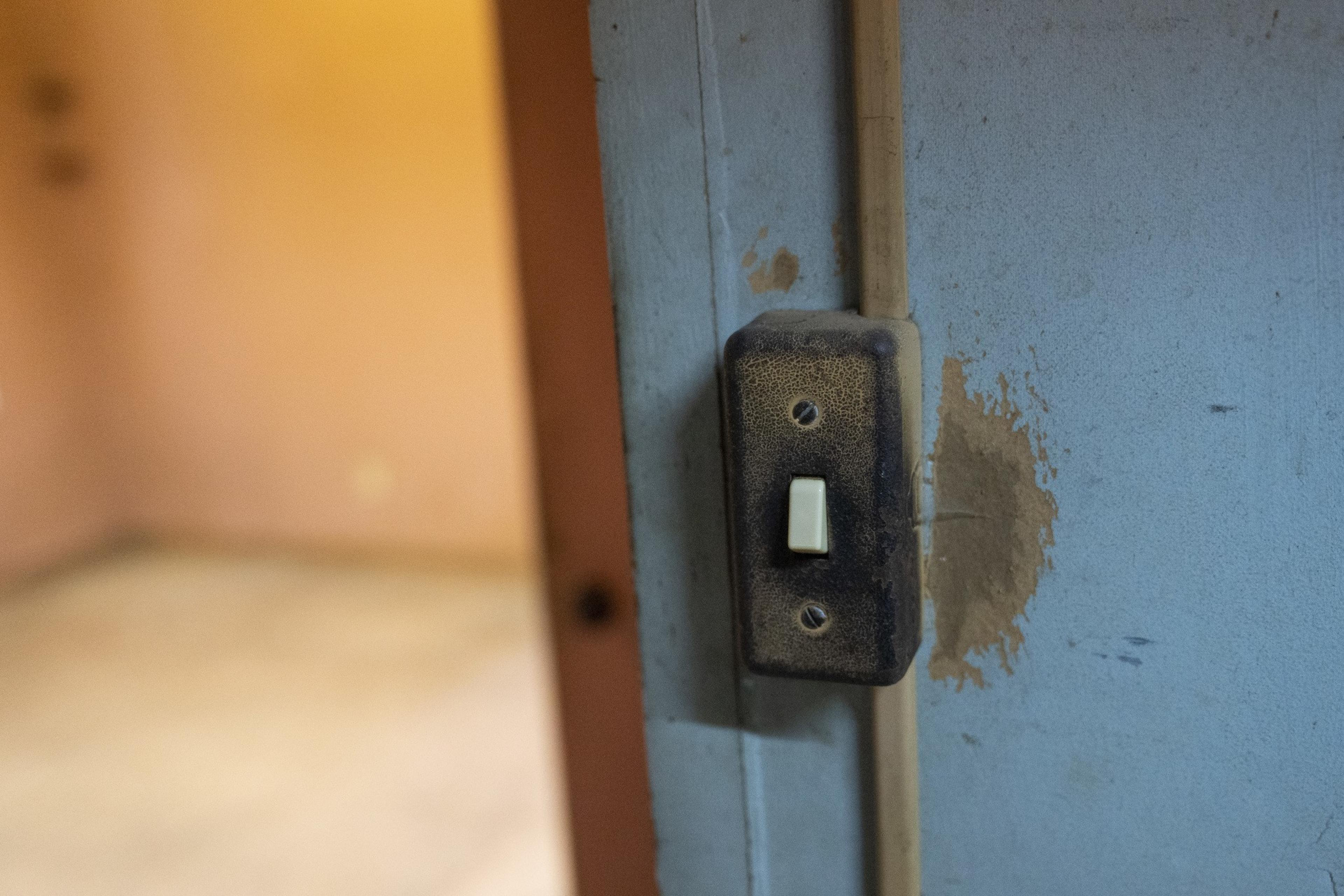A dusty, worn light switch is attached to a blue wall with peeling paint, situated near a doorway leading to a dimly lit, beige-colored room.