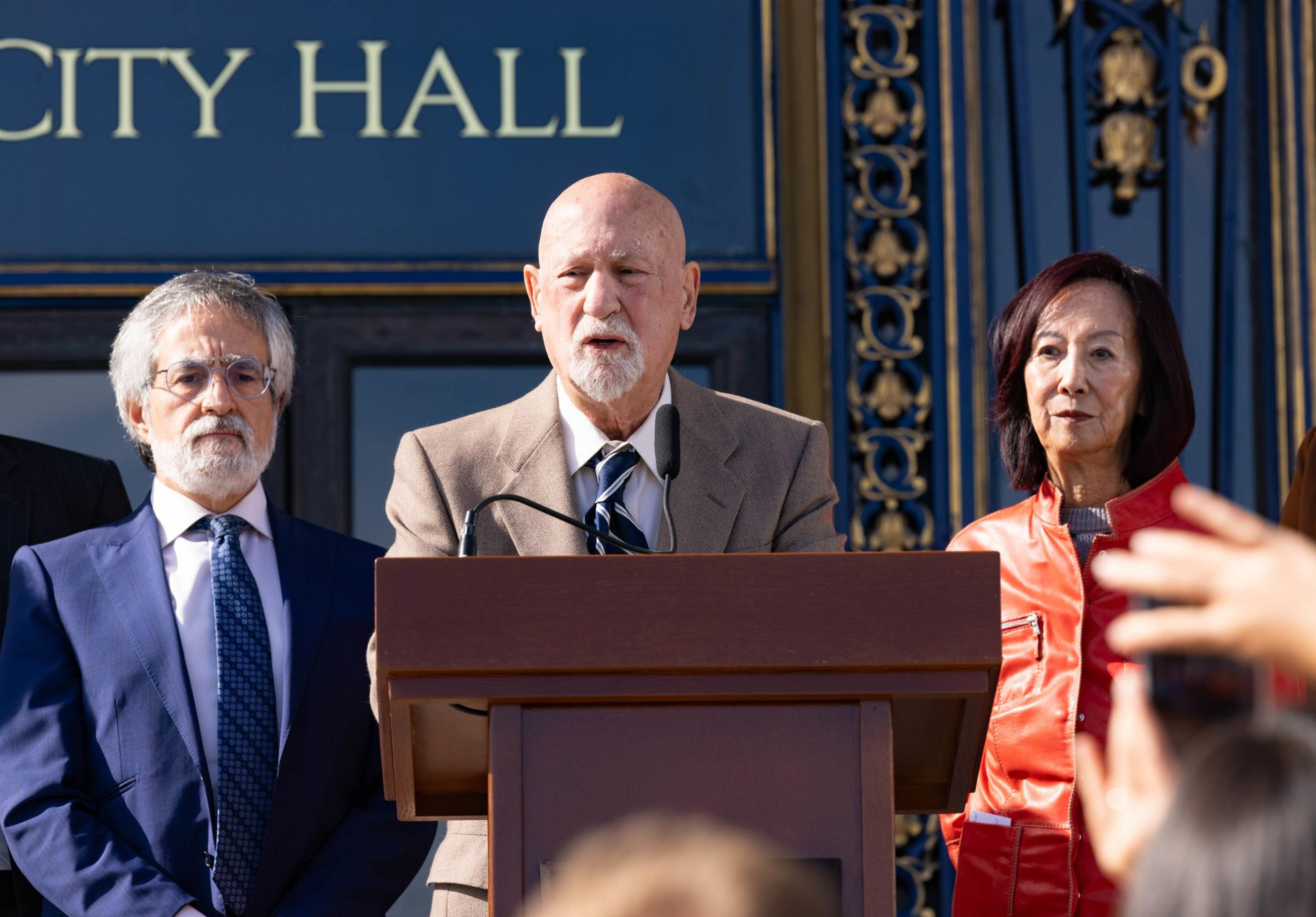 A man speaks into a microphone while standing in front of a big glass door that reads &quot;City Hall&quot; as 2 other stand beside him during a press conference.