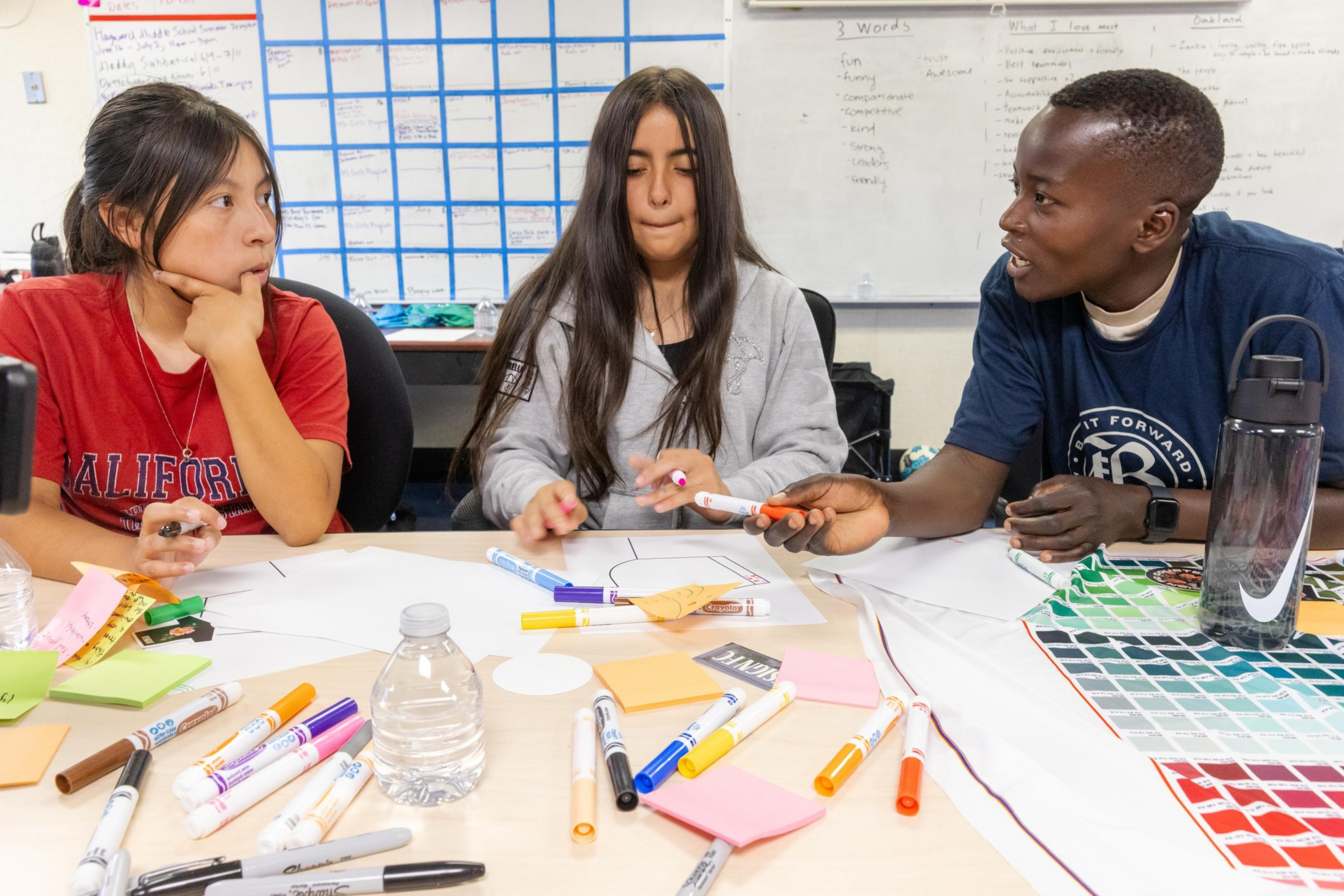 Three students sit around a table covered with markers, sticky notes, and papers, engaged in a discussion in a classroom.