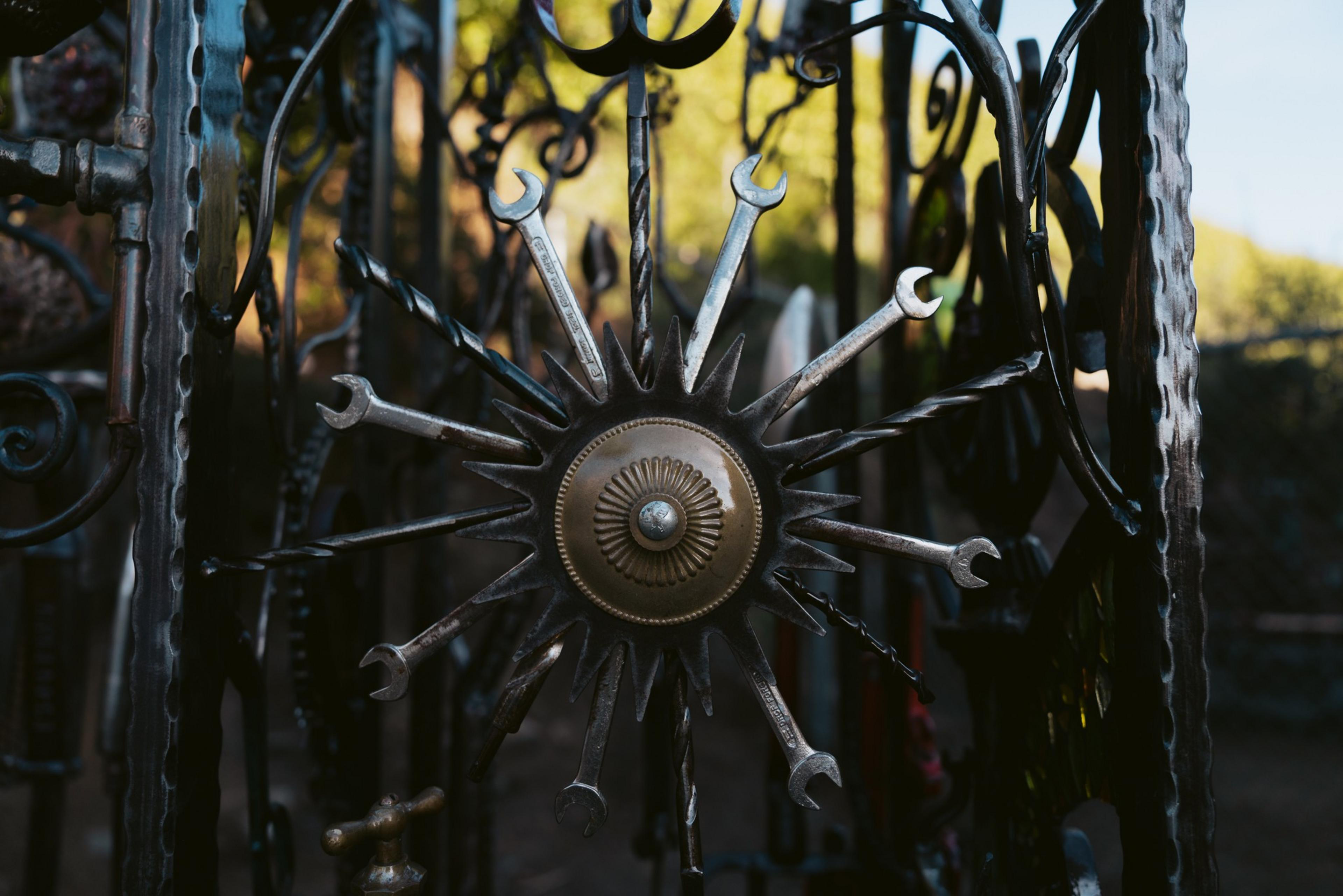 A metal gate features a sun-shaped design with wrenches as rays and a decorative circular center, set against a blurred outdoor background.