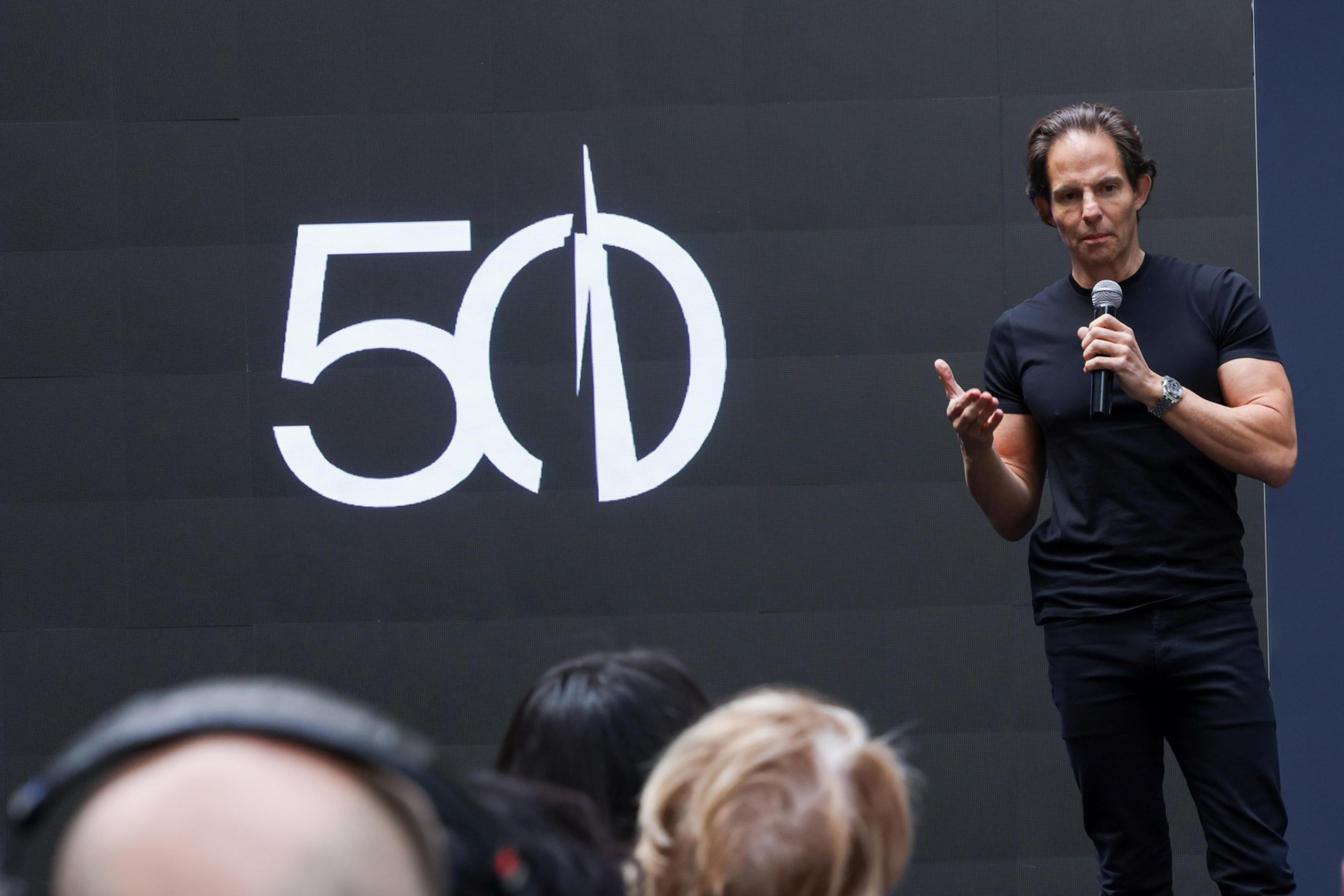 A man in a black shirt is speaking into a microphone on a stage with a black background displaying a large white "50" and stylized "D" behind him.