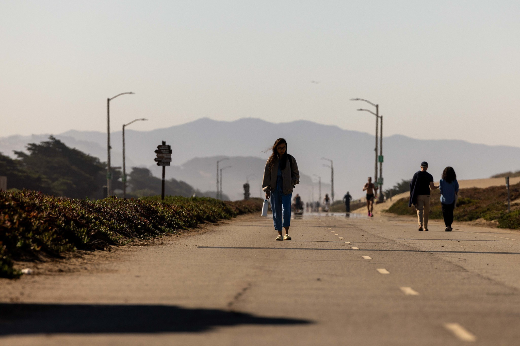 A few people walk along a paved path with mountains in the background under clear skies and streetlights lining the path.
