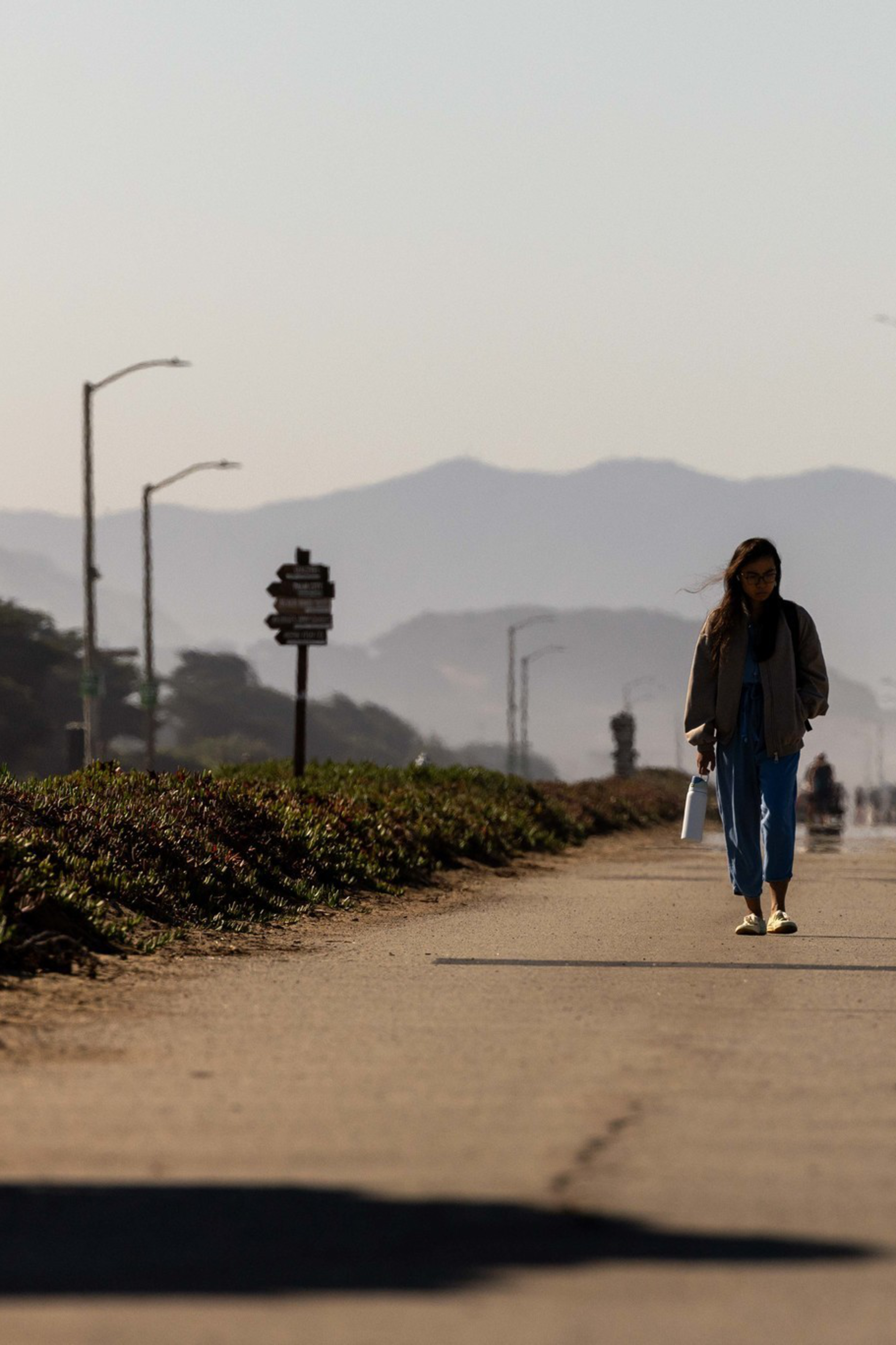 A few people walk along a paved path with mountains in the background under clear skies and streetlights lining the path.