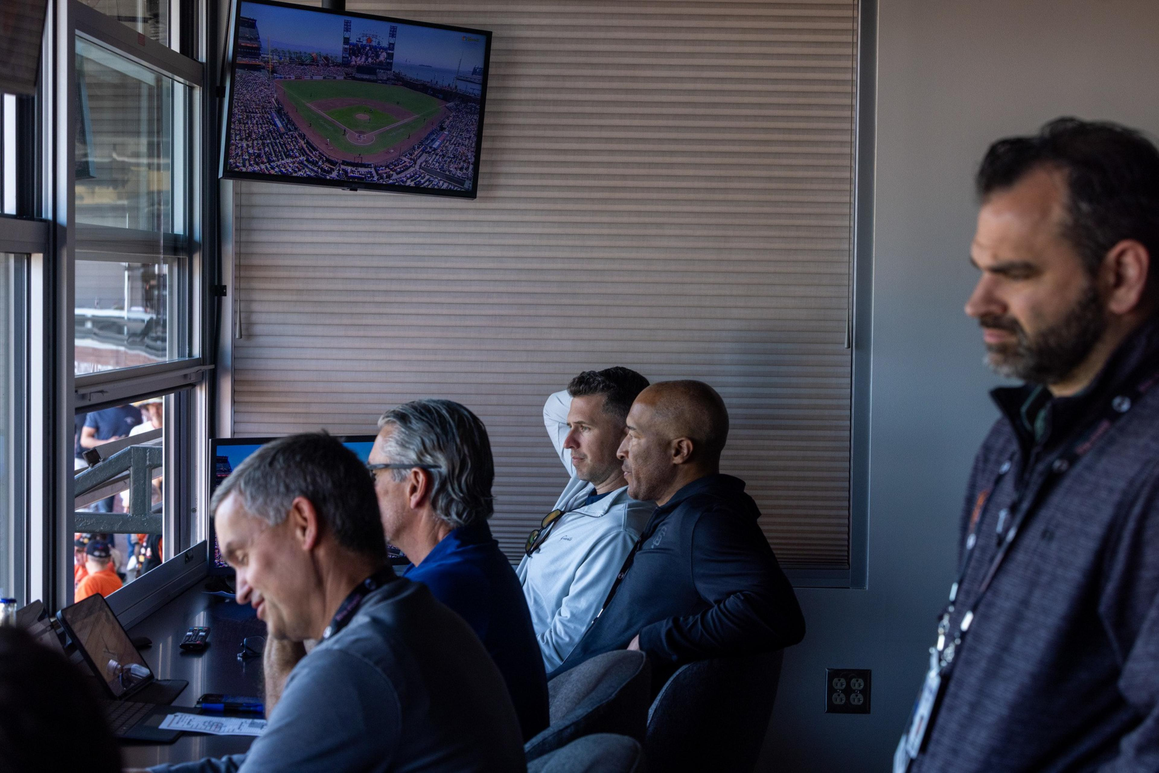 Several men are seated in a room watching a baseball game on screens. A window is open to the field, and a small TV shows the game. They appear focused.