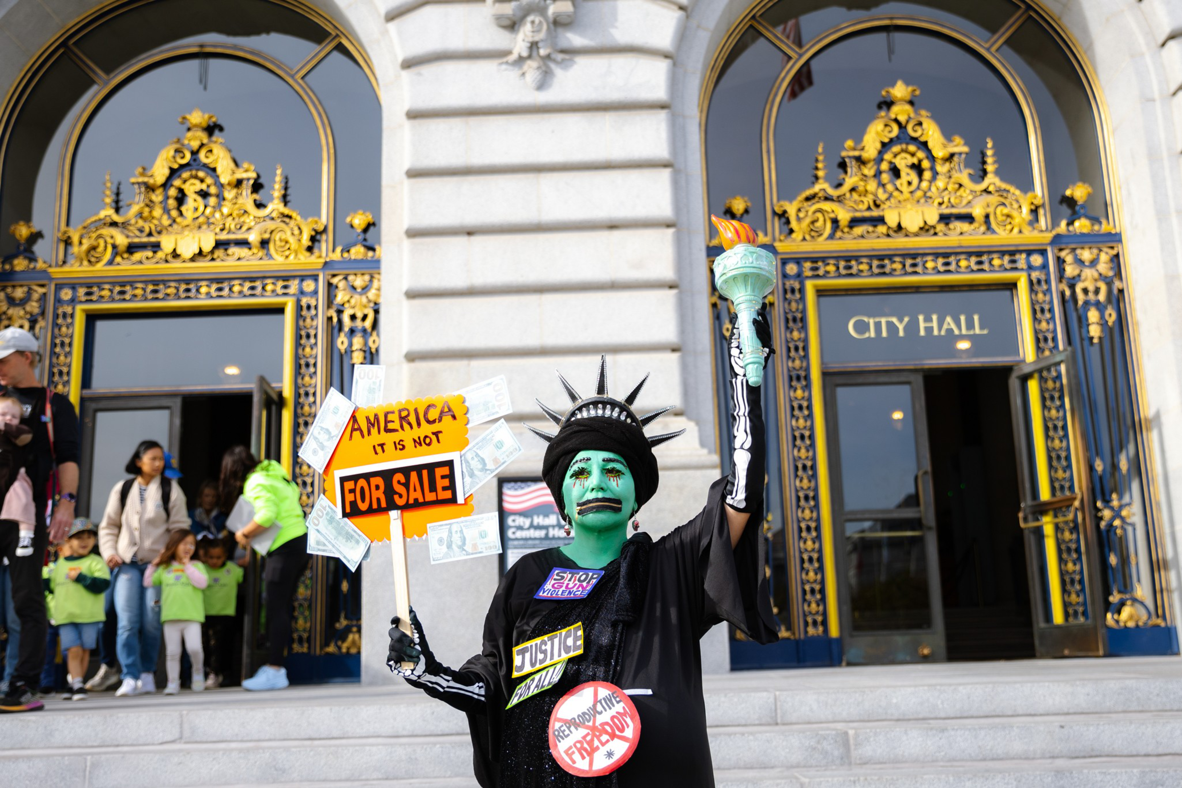 A person dressed as the Statue of Liberty holds a torch and a sign reading &quot;America It Is Not For Sale&quot; on the steps of a grand city hall entrance.