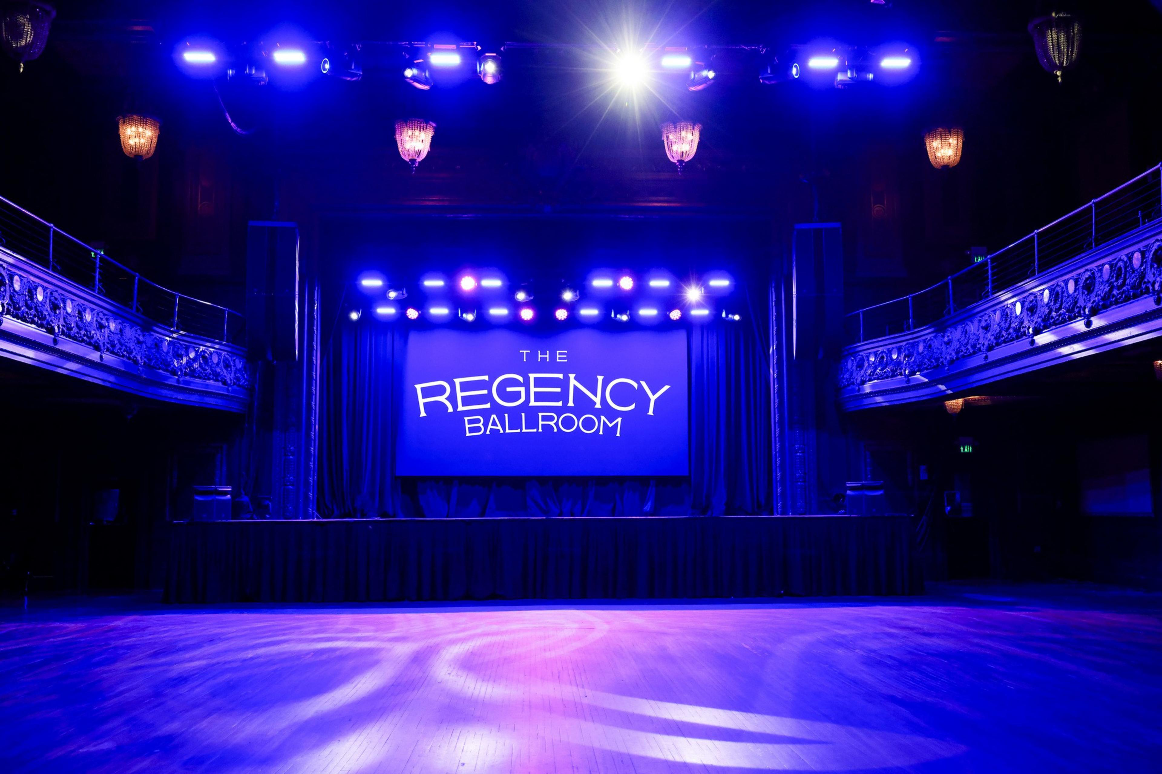 A ballroom with a wooden floor is dimly lit in blue and purple. A stage at the back features a screen displaying "The Regency Ballroom." Five chandeliers hang above.