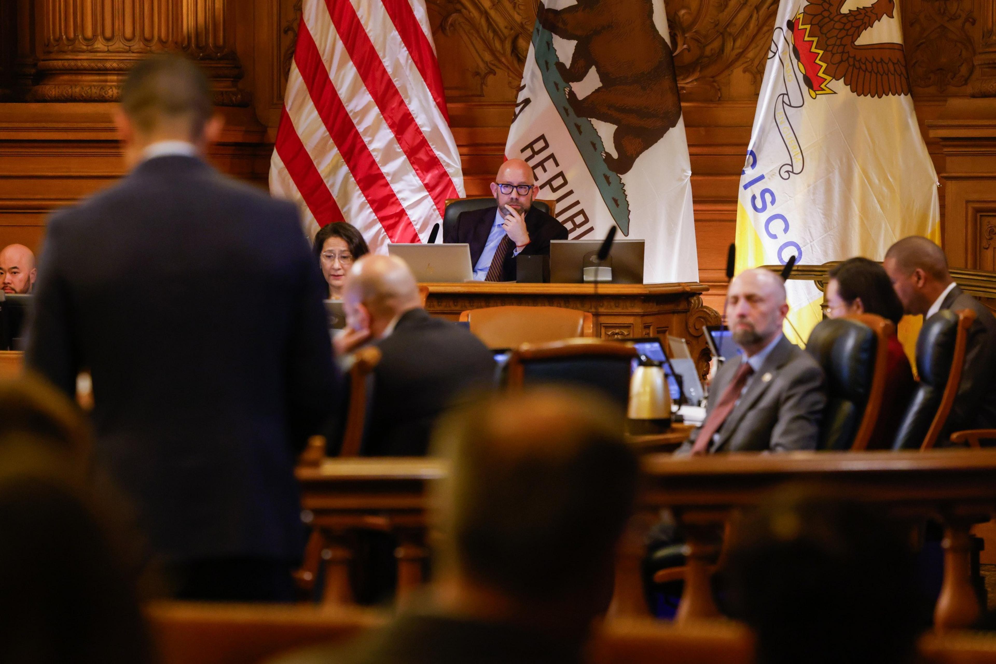 A person sits at a desk with laptops and microphones, surrounded by others in a formal room. American, California, and San Francisco flags are on display.