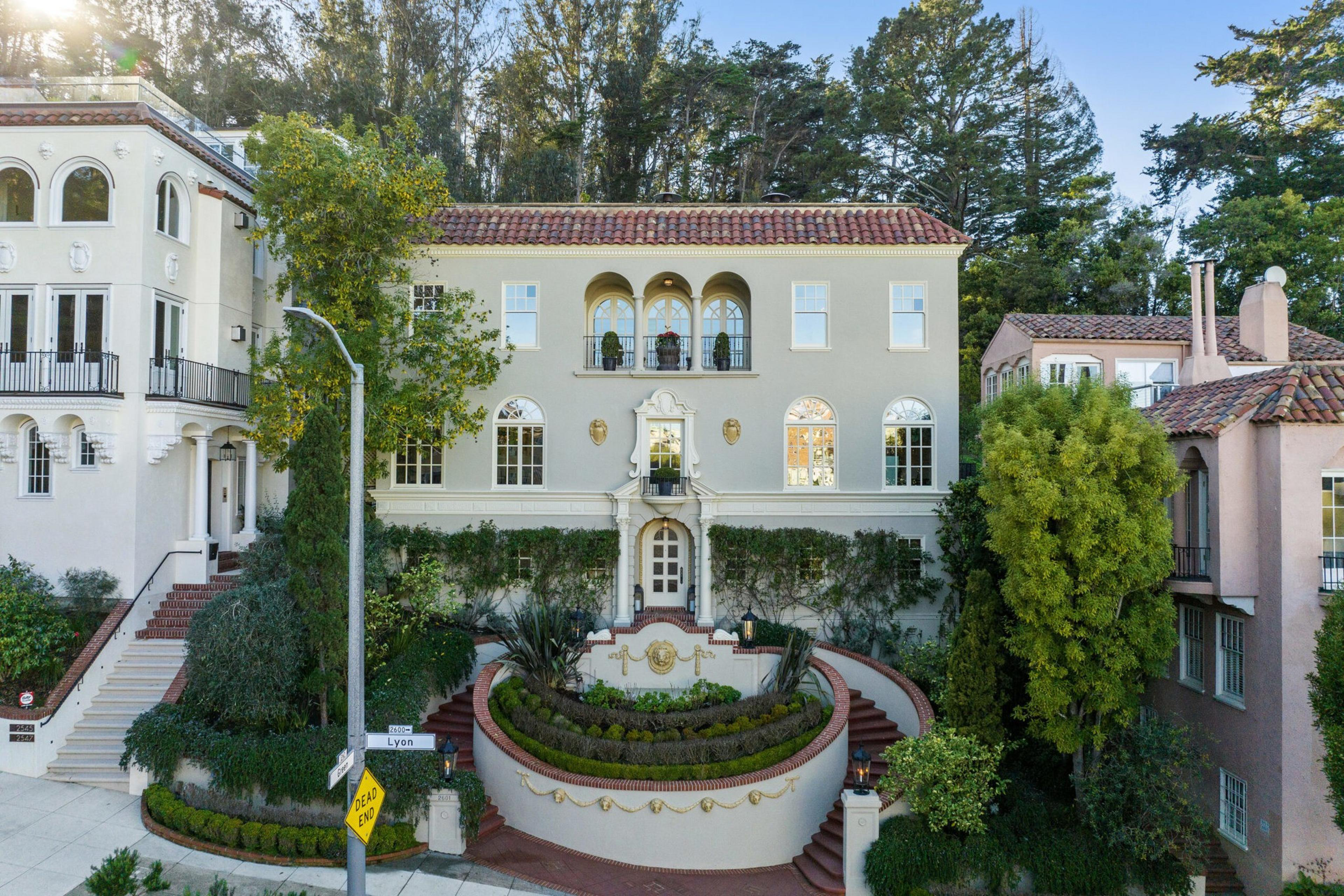 A large, elegant Mediterranean-style house with arched windows, a tiled roof, circular front garden, and two curved staircases leading to the entrance.