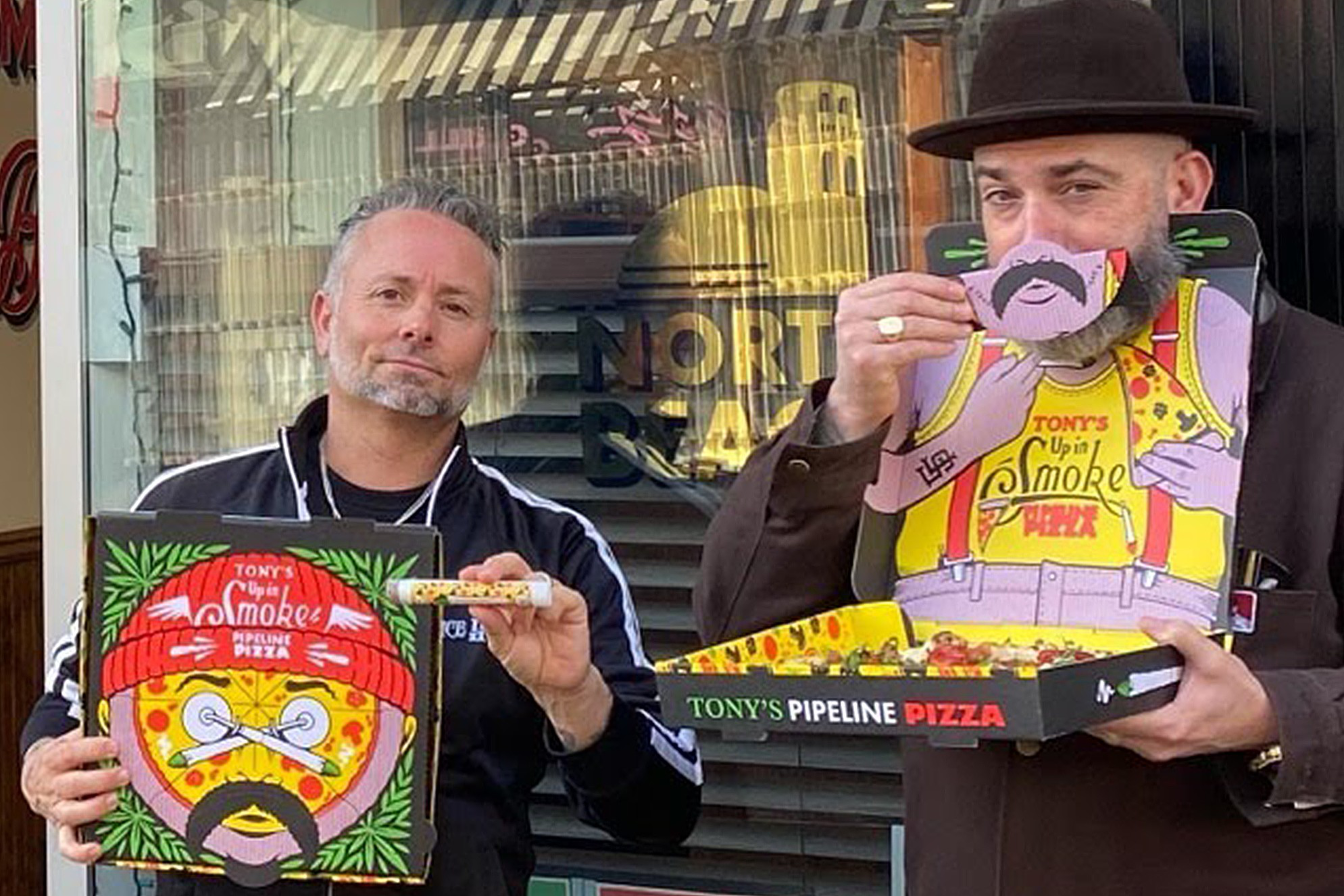 Two men stand outside holding pizza boxes from Tony's Pipeline Pizza; one man holds a slice, the other wears a hat and holds a cutout with a drawn mustache.