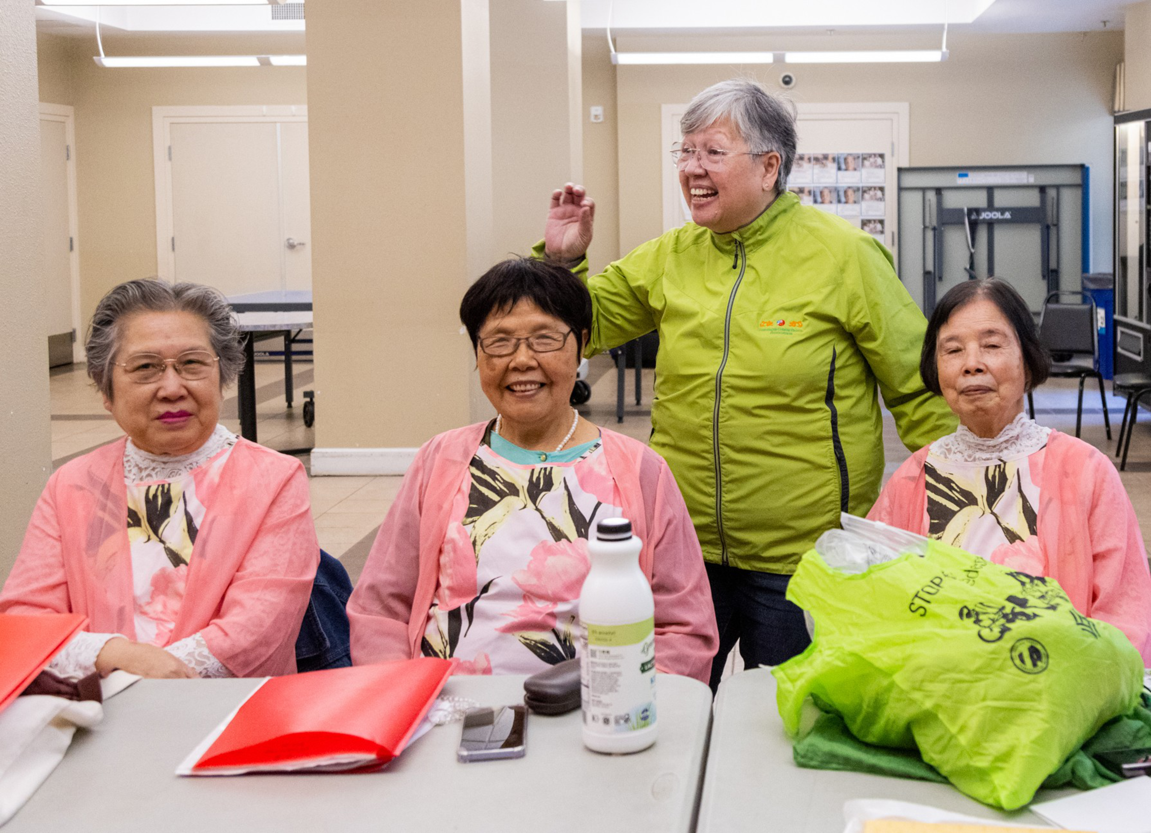 Four senior women in a community room, three seated and wearing pink smocks, one standing in a green jacket, all appear cheerful.