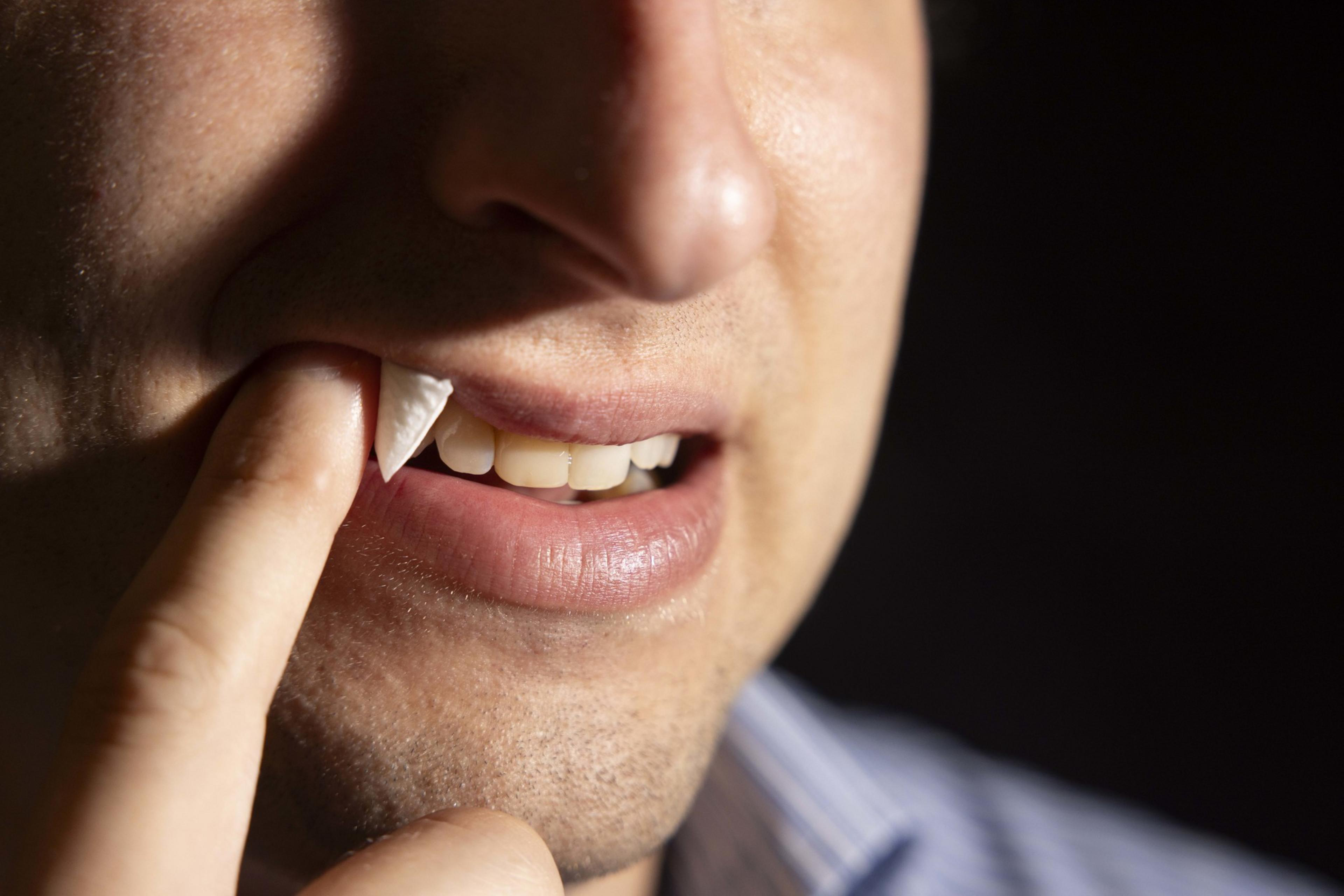 A person is using their finger to hold a fake vampire fang to their upper teeth, showing part of their face with a focus on their mouth and the white, pointed fang.