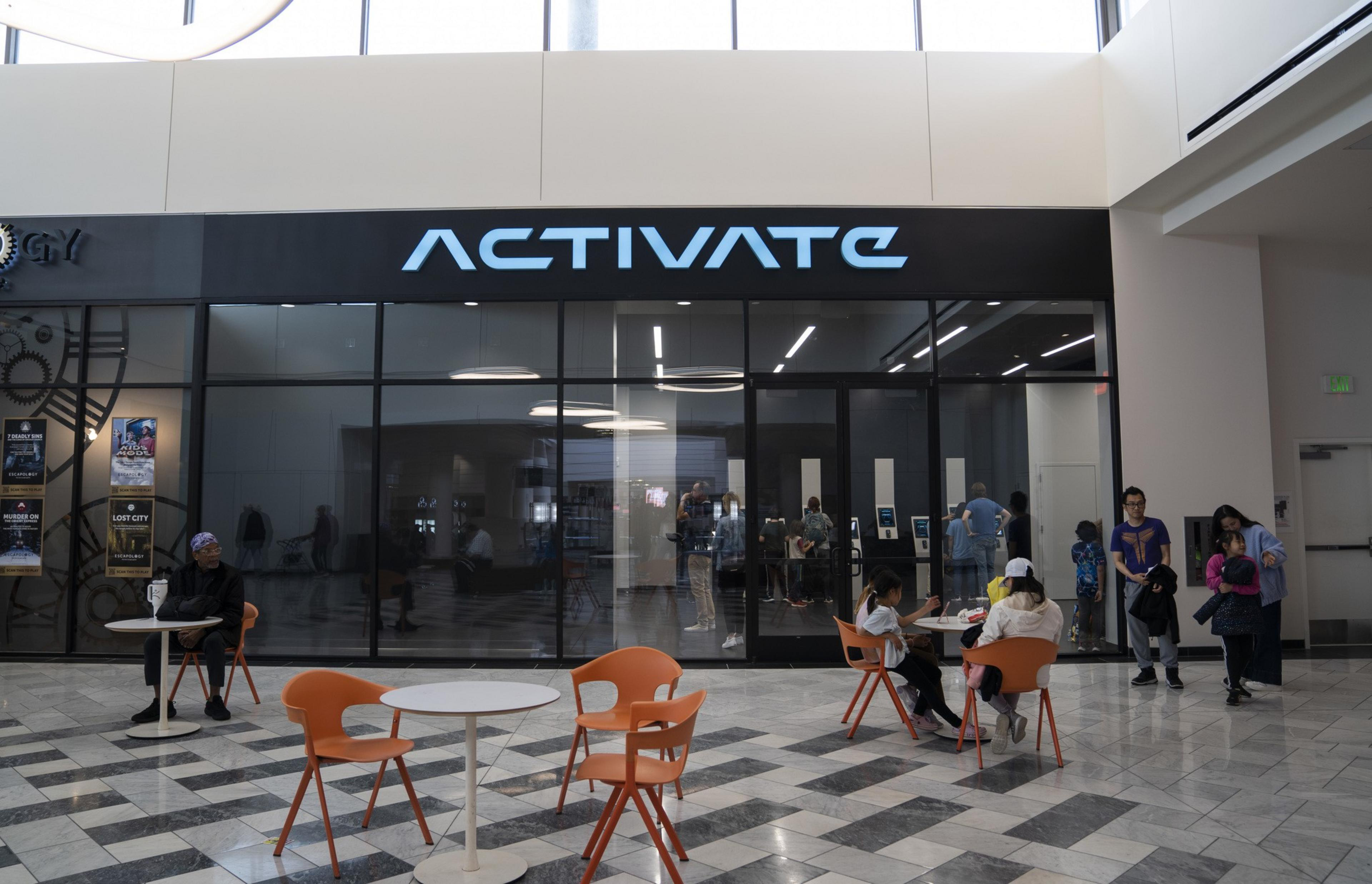 The image shows a store called &quot;ACTIVATE&quot; with a glass front, bright interior lighting, and people inside. In front, there are orange chairs and tables with people sitting.