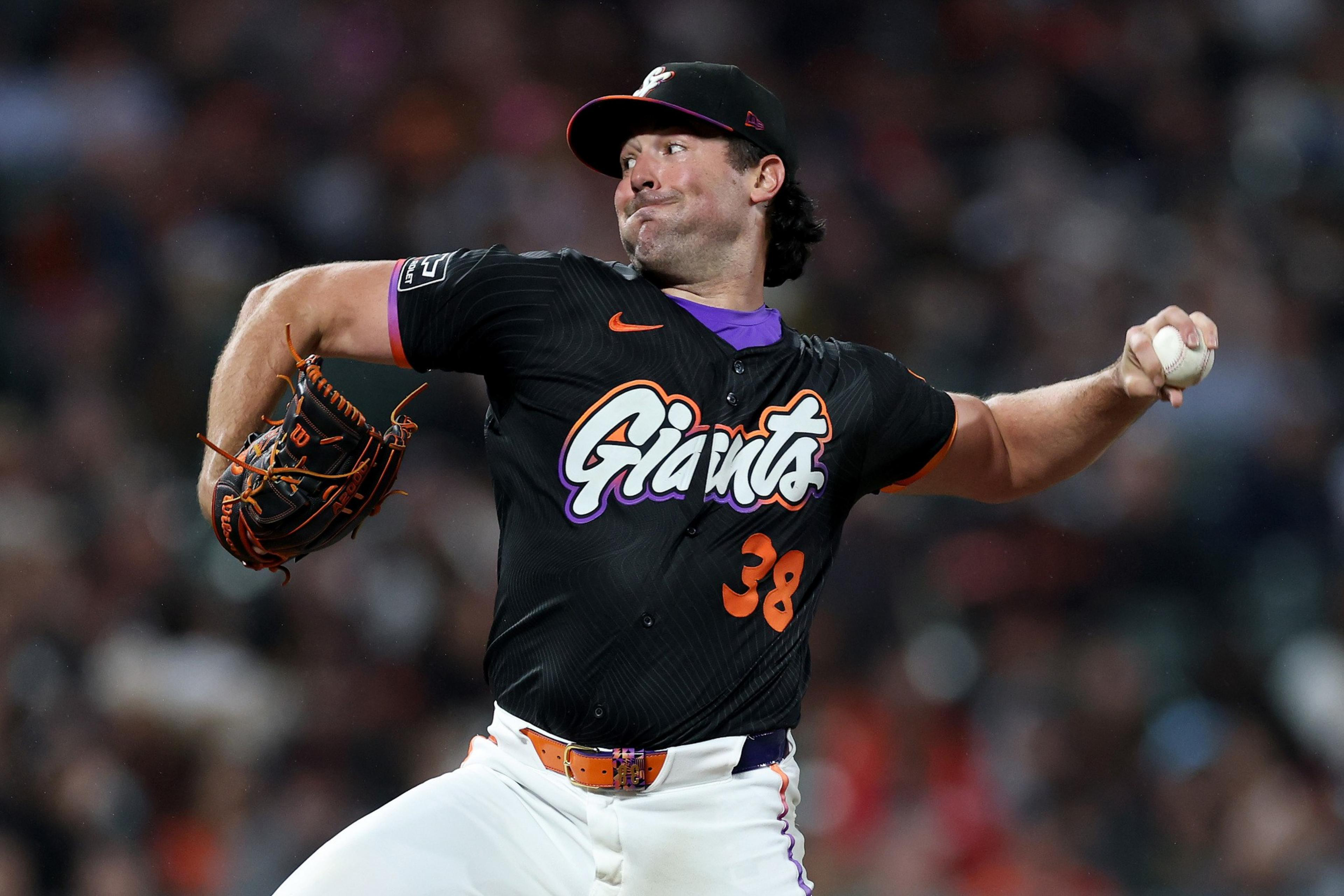 A baseball pitcher in a black Giants jersey with number 38 is mid-throw, gripping a ball in his right hand and wearing a glove on his left.