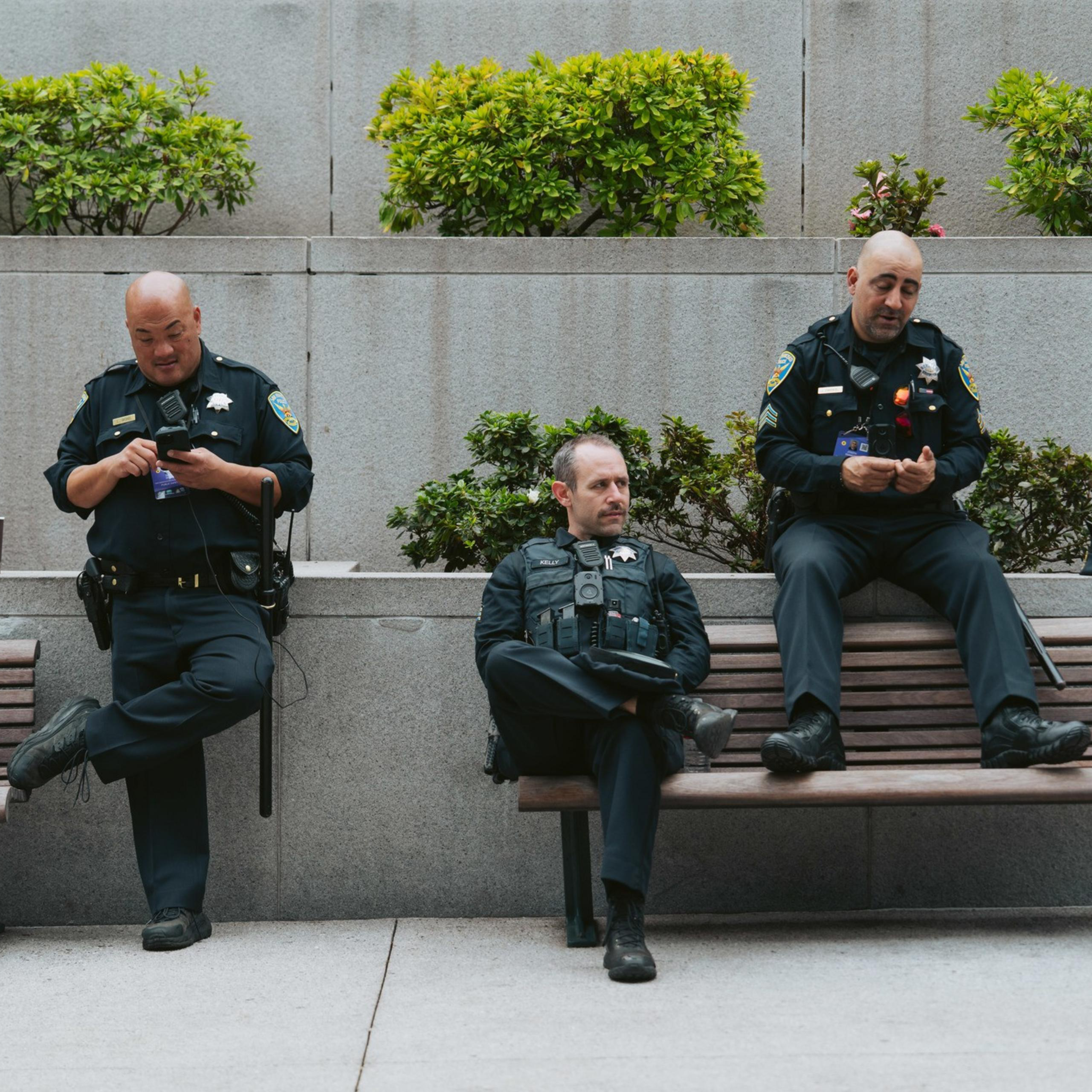 Three police officers are sitting on a bench; two are on their phones, and one looks pensive. There's greenery behind them on a gray wall.