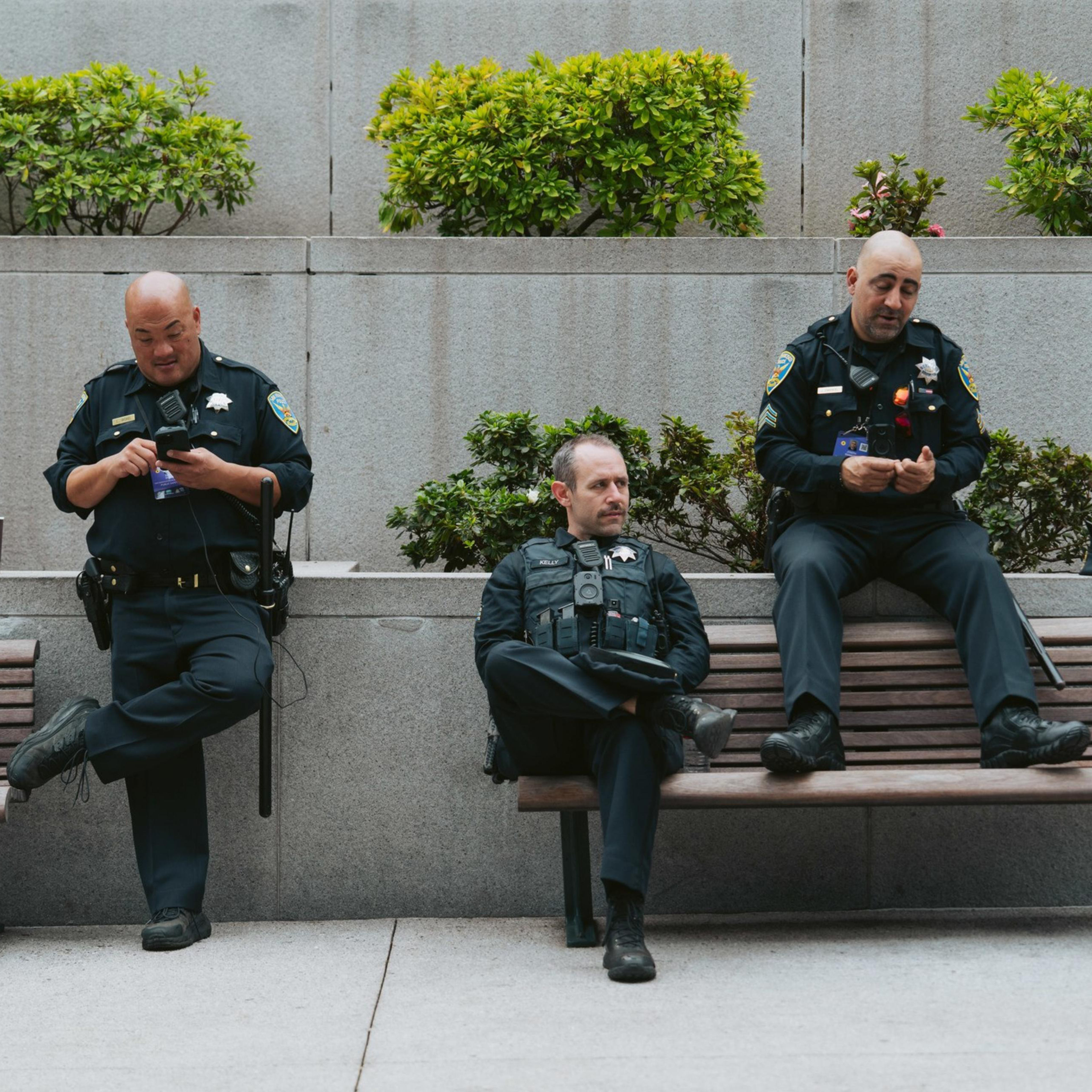 Three police officers are sitting on a bench; two are on their phones, and one looks pensive. There's greenery behind them on a gray wall.