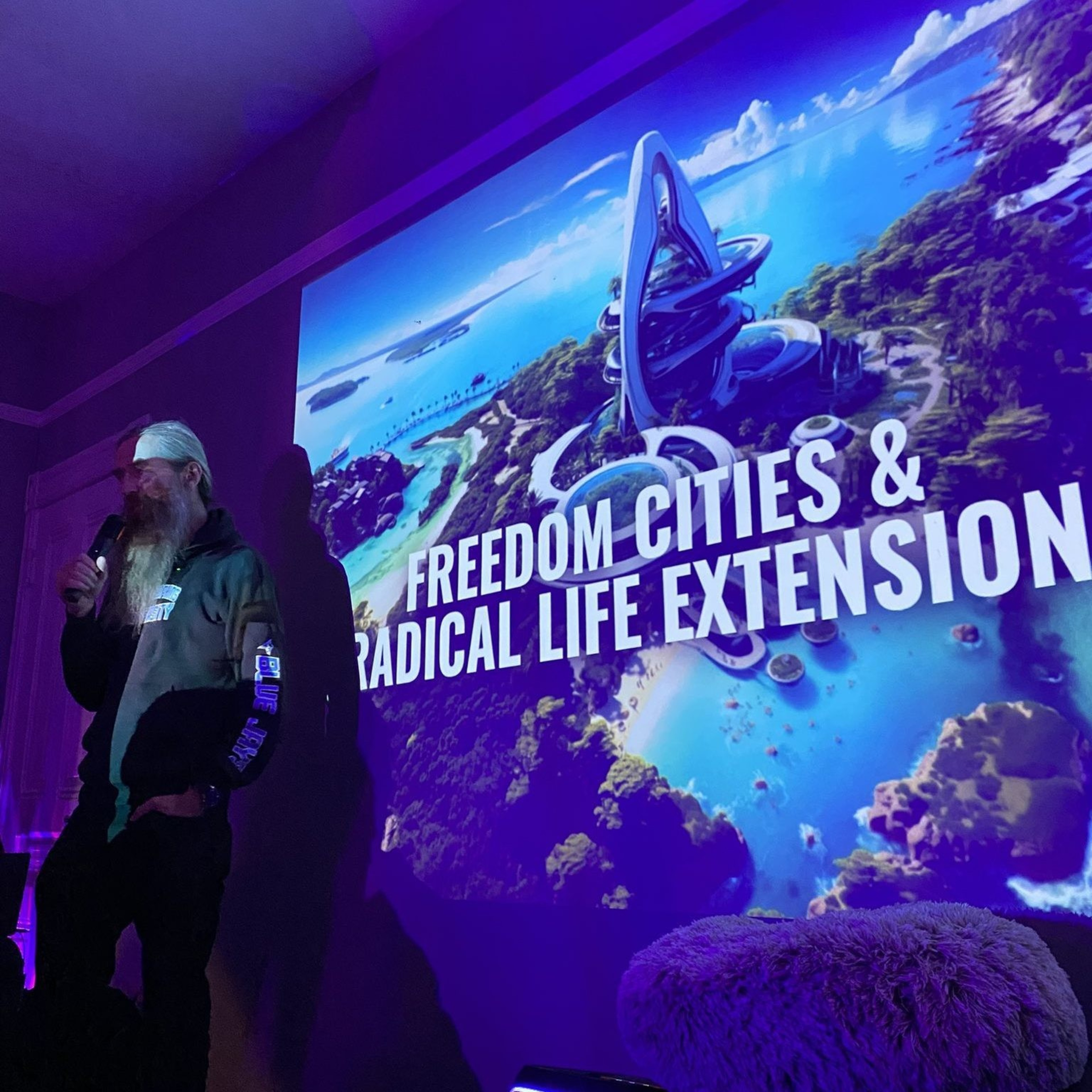 A person stands in dim lighting, speaking into a microphone. To their right, a vibrant image of a futuristic city is projected, labeled &quot;Freedom Cities &amp; Radical Life Extension.&quot;