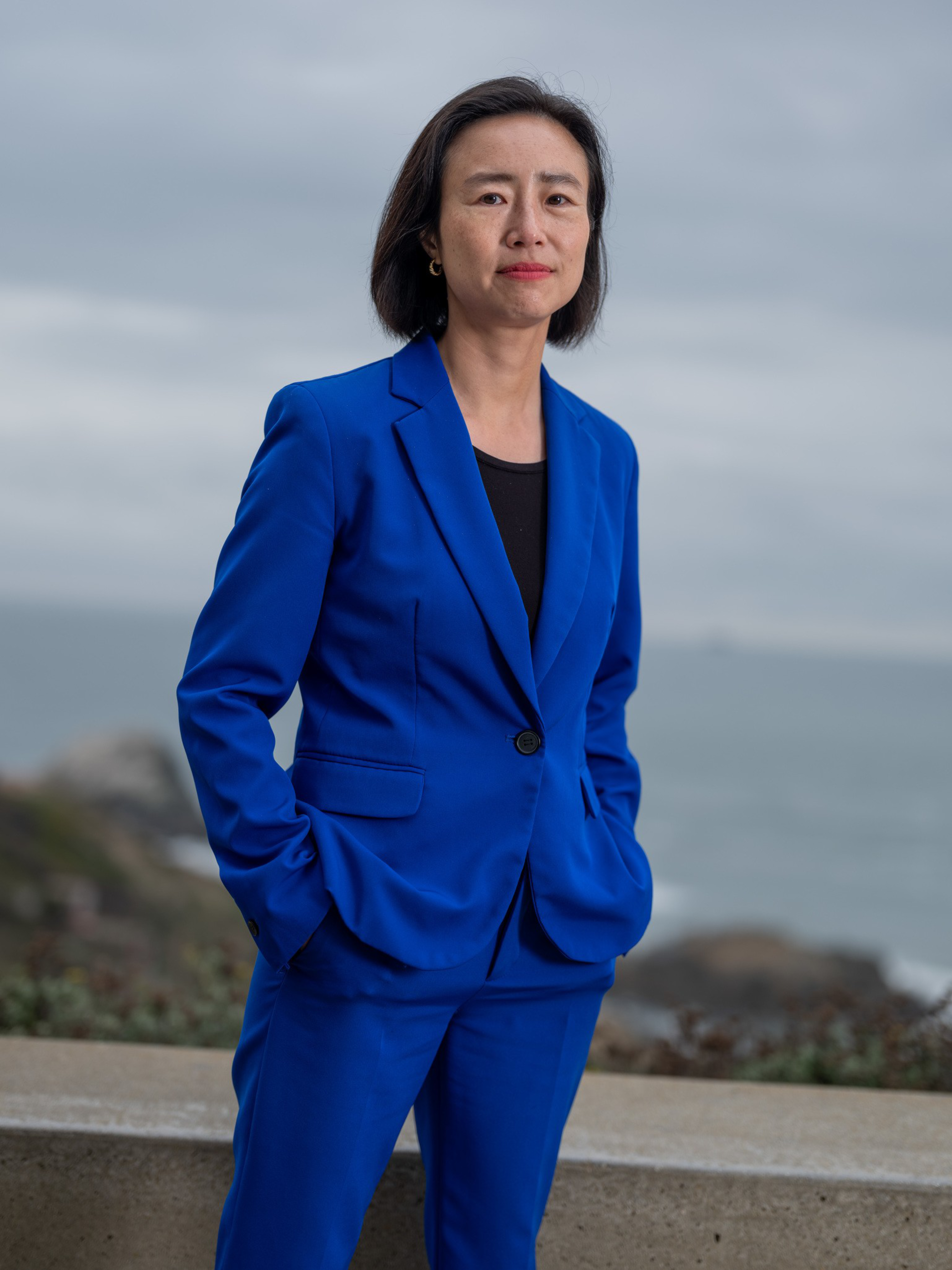 A woman in a bright blue suit with hands in pockets stands outdoors, with a calm expression and a cloudy sky over a body of water in the background.