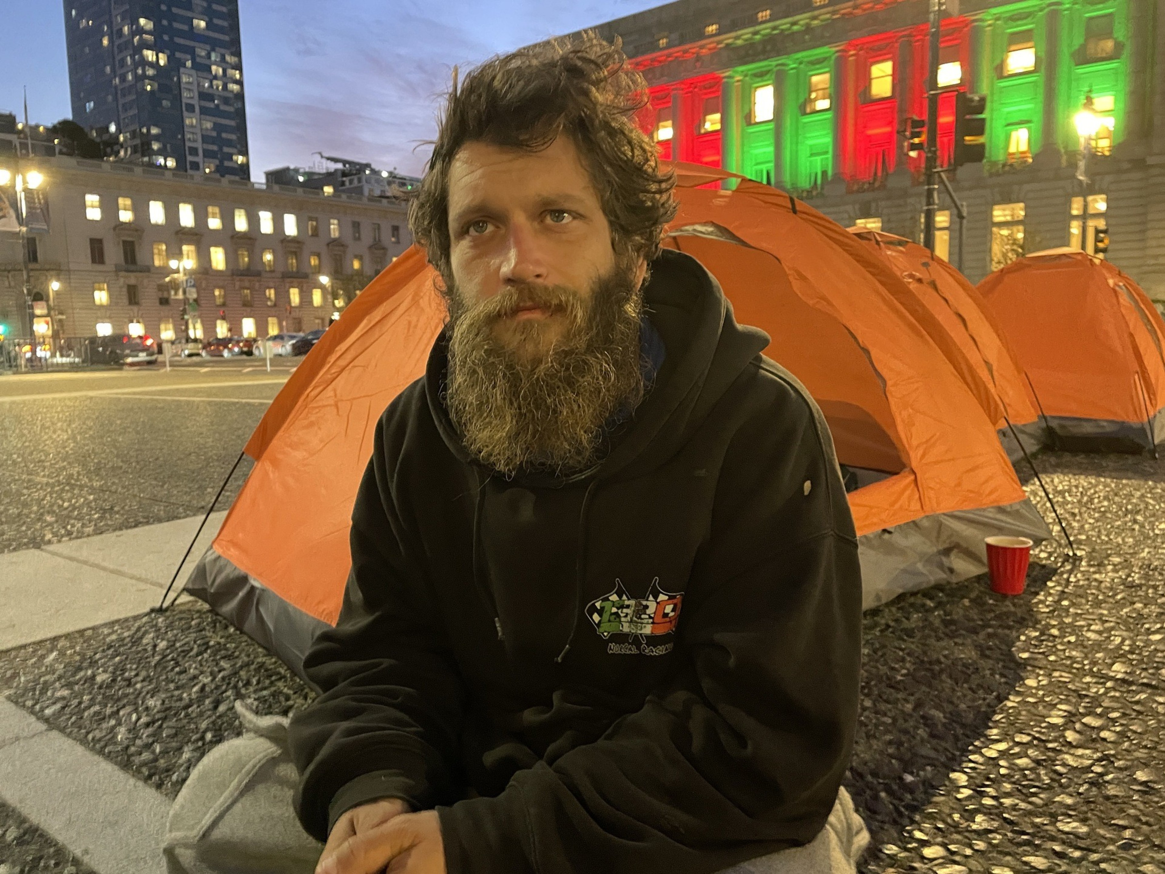 A bearded man in a black hoodie sits outside an orange tent in a lit city square at dusk. Buildings with illuminated windows and colorful lights are in the background.