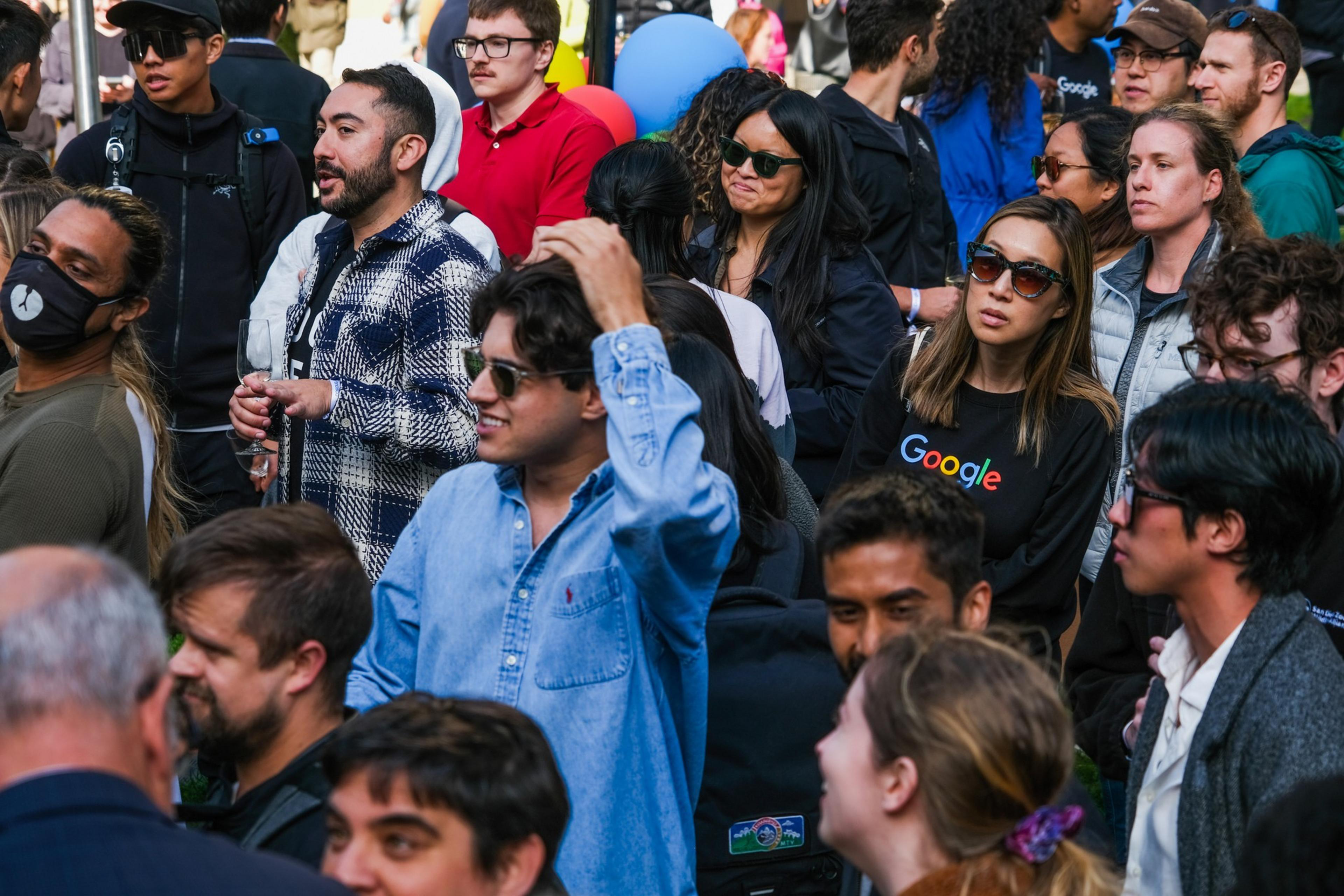 A crowd of diverse people, many wearing sunglasses and casual clothing, including some with Google logos. One person holds a wine glass, and a large blue balloon is visible.