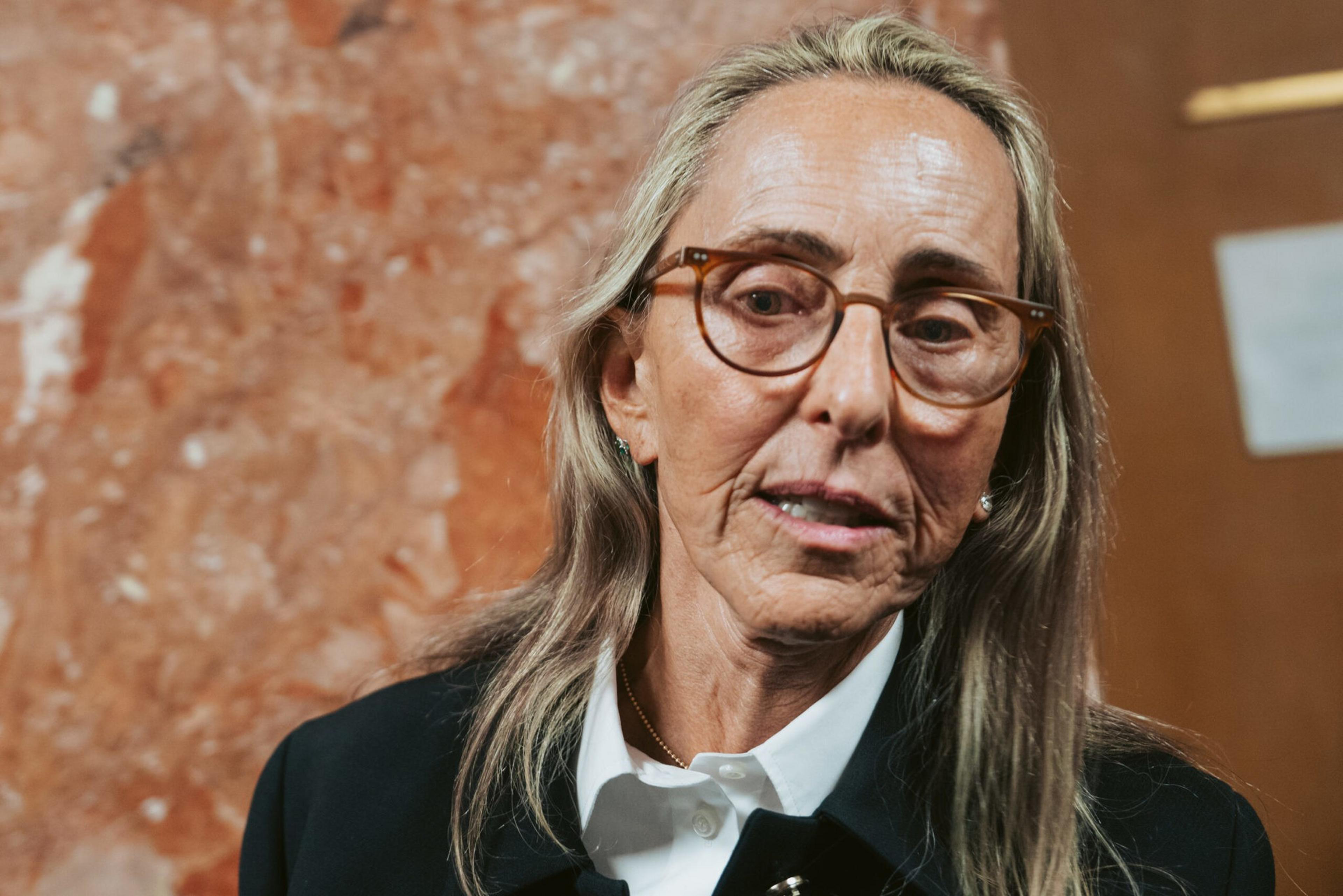 An older woman with long gray hair wears tortoiseshell glasses, a white collared shirt, and a dark jacket, standing against a reddish-brown textured background.