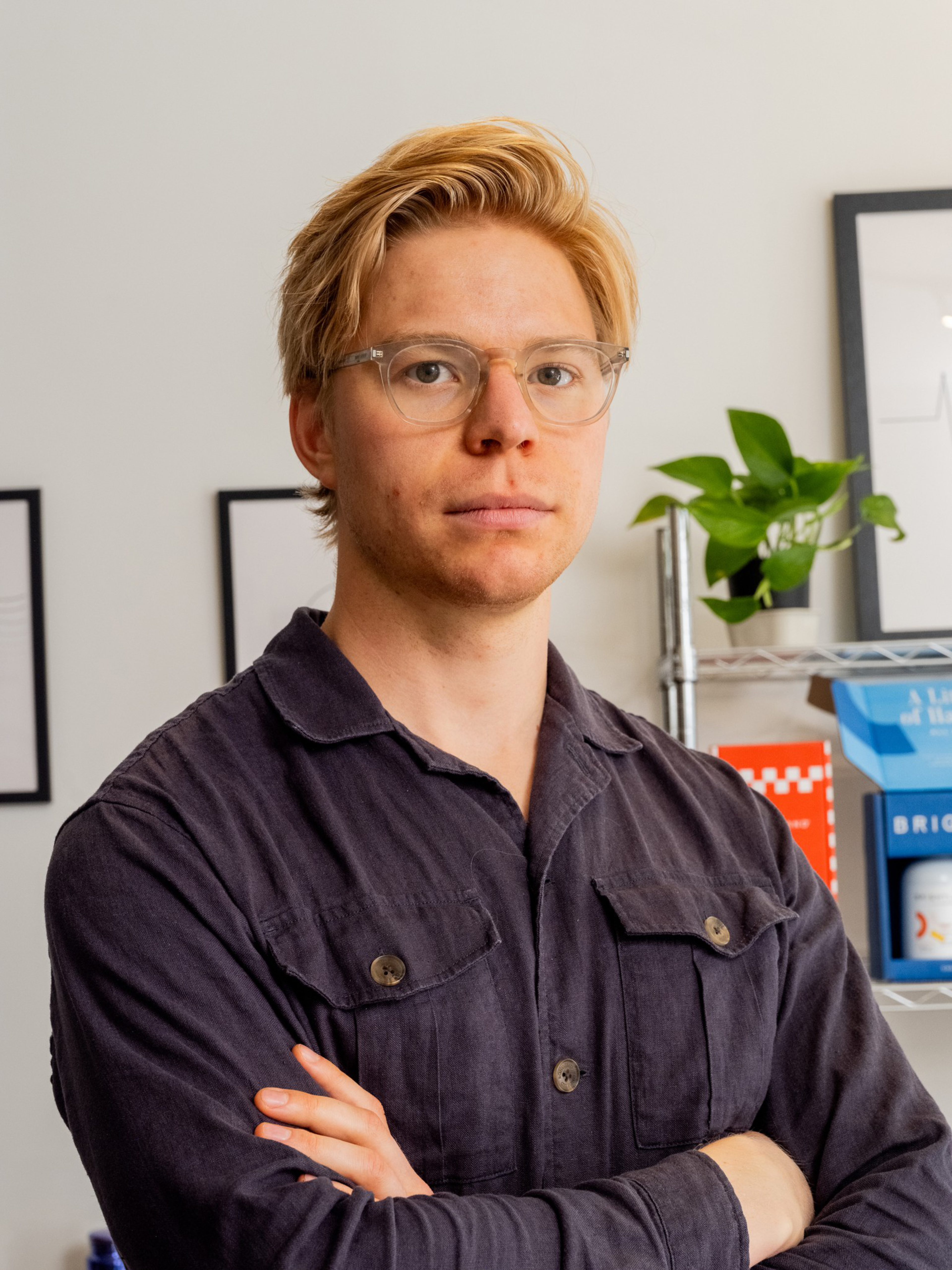 A young person with blonde hair and clear glasses wears a dark shirt, standing with arms crossed inside a room with framed art and a green plant.