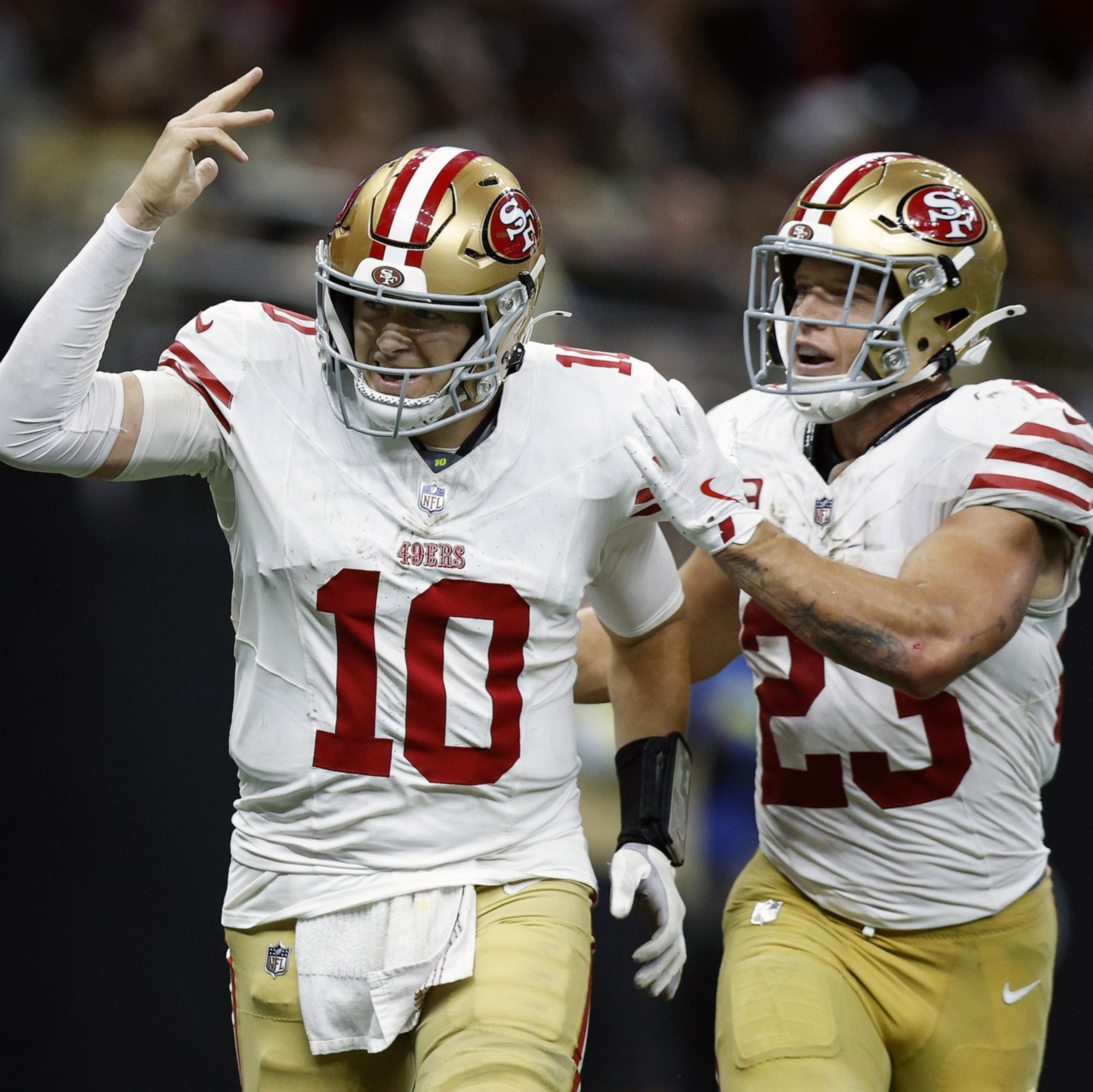 Two San Francisco 49ers players in white jerseys and gold helmets celebrate, with one raising his hand and the other reaching out to him.