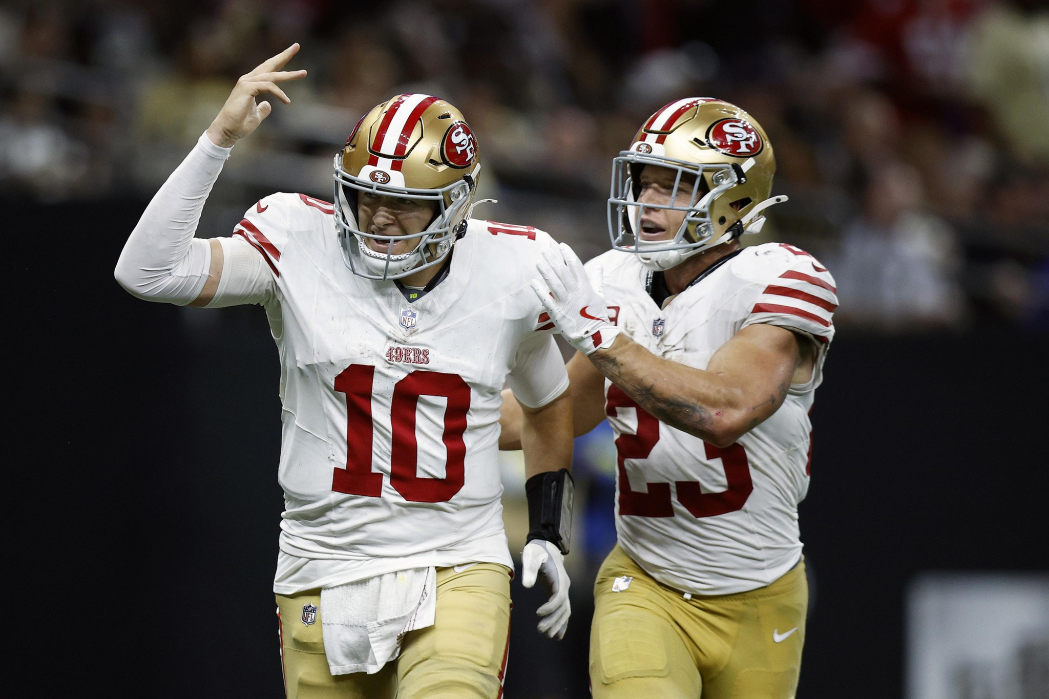 Two San Francisco 49ers players in white jerseys and gold helmets celebrate, with one raising his hand and the other reaching out to him.