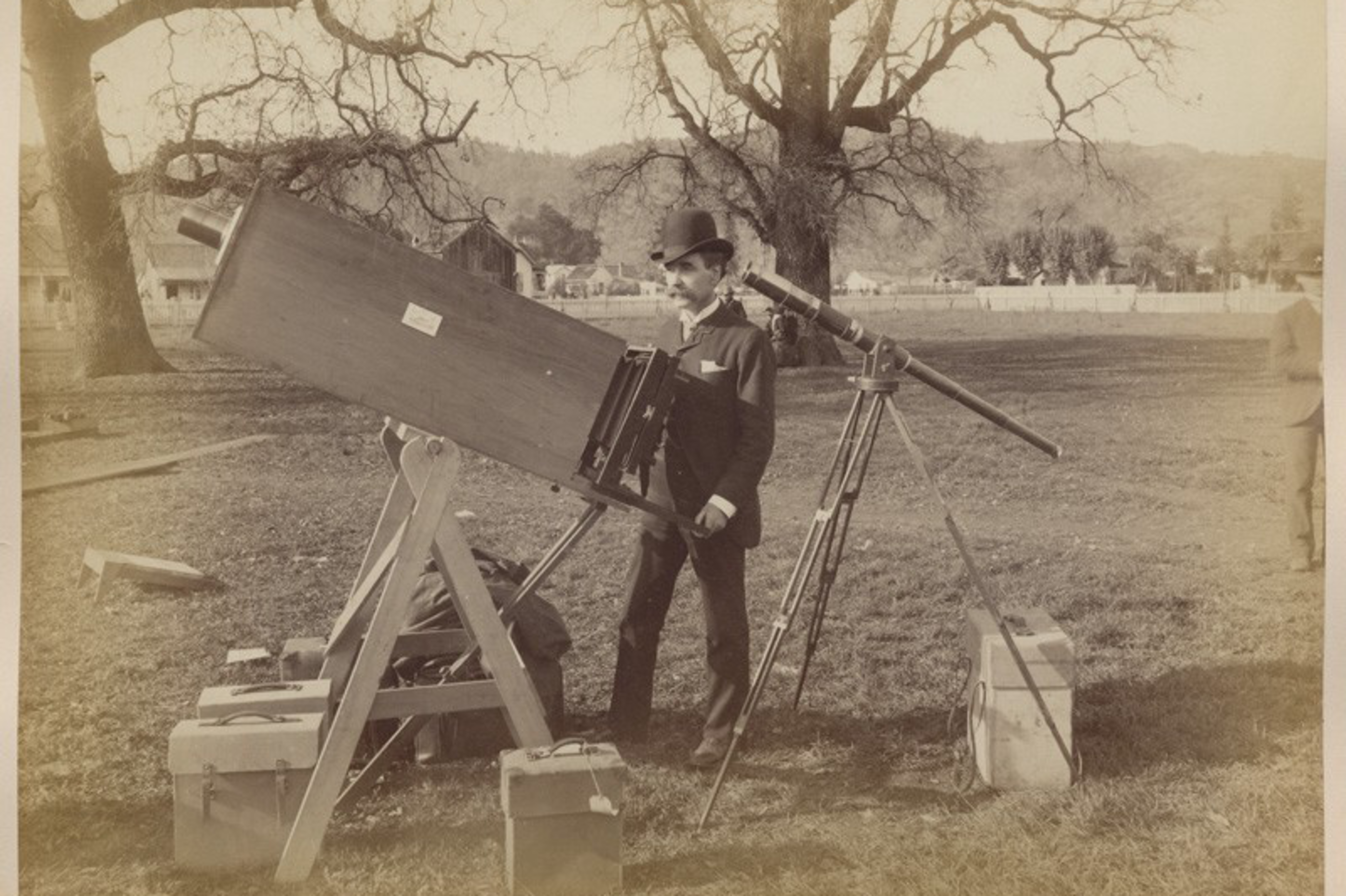 A man in vintage attire operates a large telescope outdoors; boxes and a leafless tree nearby.
