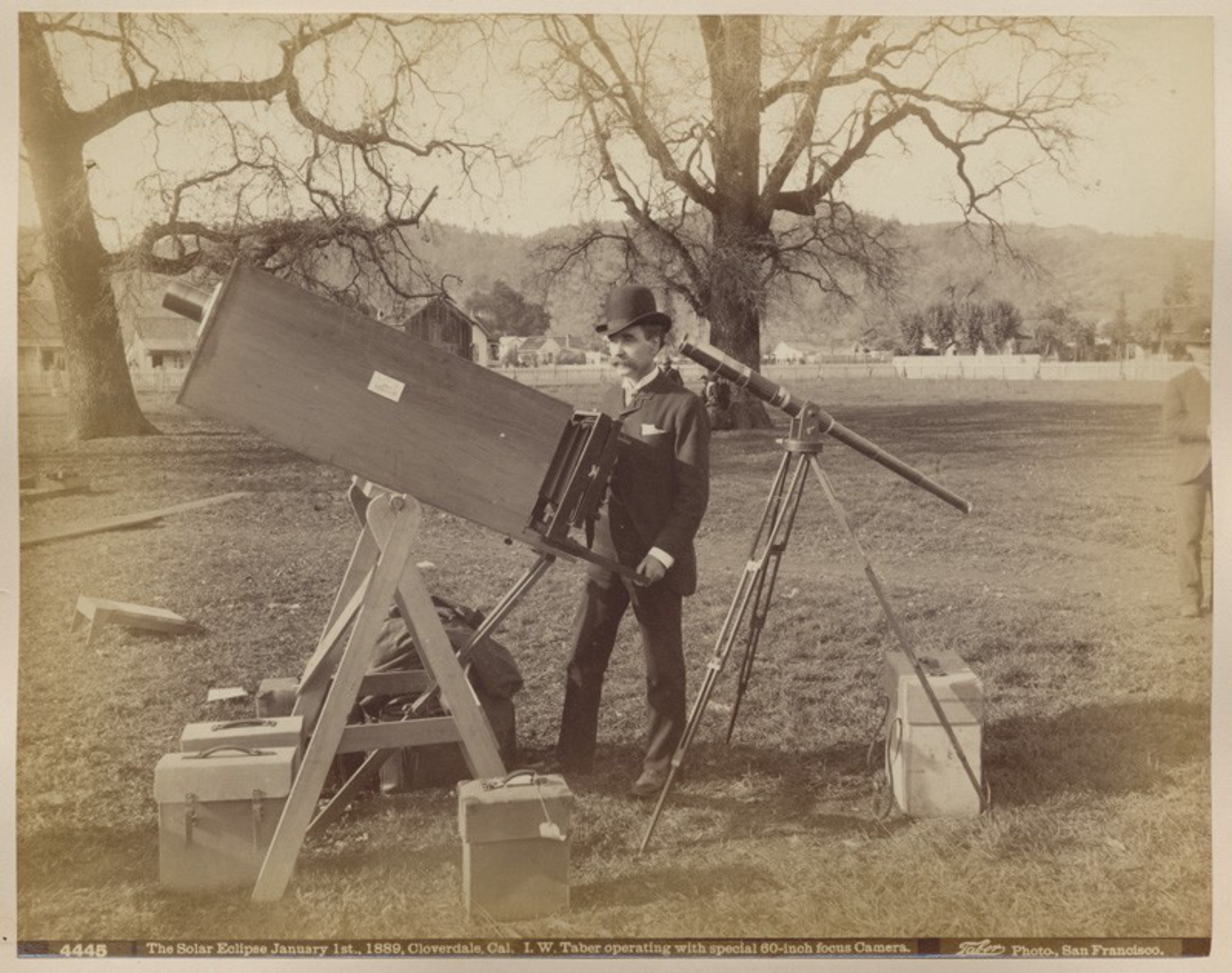 A man in vintage attire operates a large telescope outdoors; boxes and a leafless tree nearby.