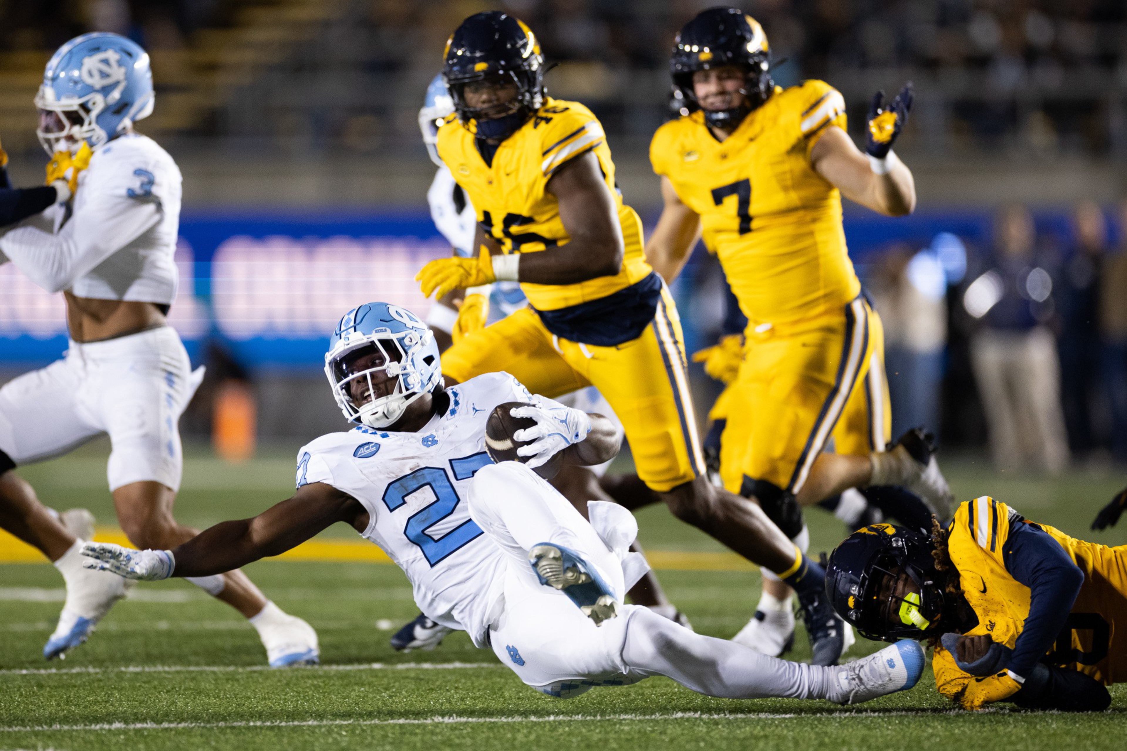 A football player in white with number 27 dives forward, holding the ball as players in yellow attempt to tackle him.