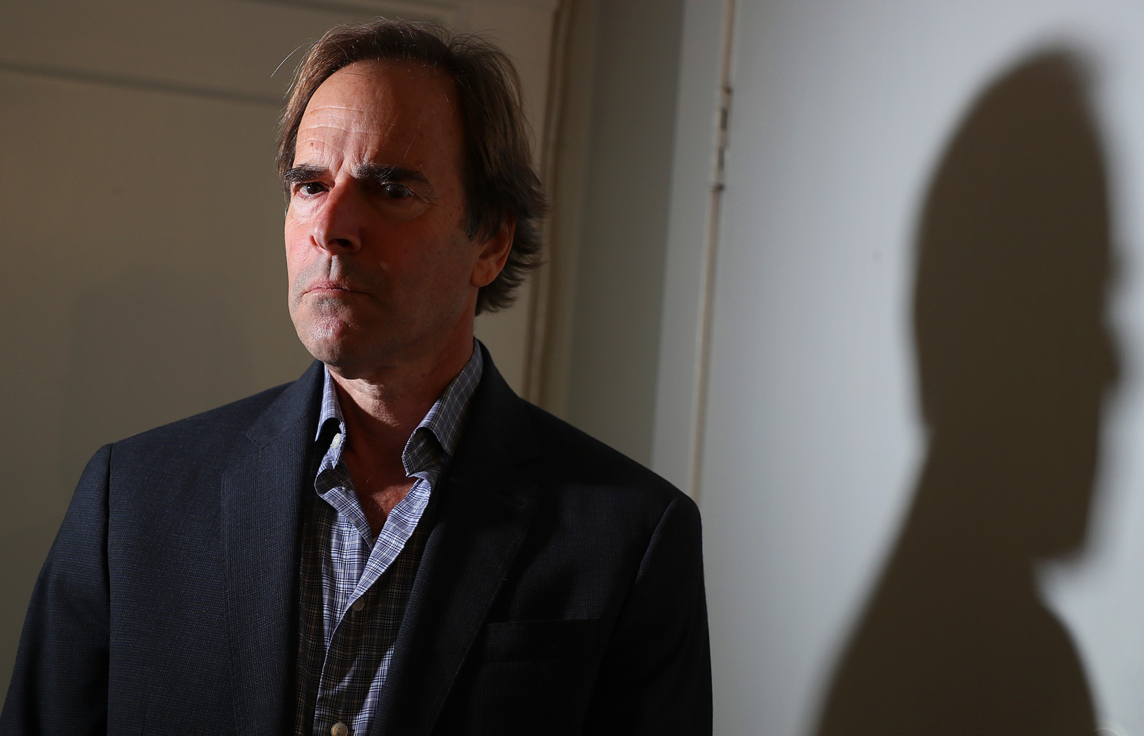 A serious-looking middle-aged man in a dark blazer and checked shirt stands against a wall, casting a dark shadow behind him.