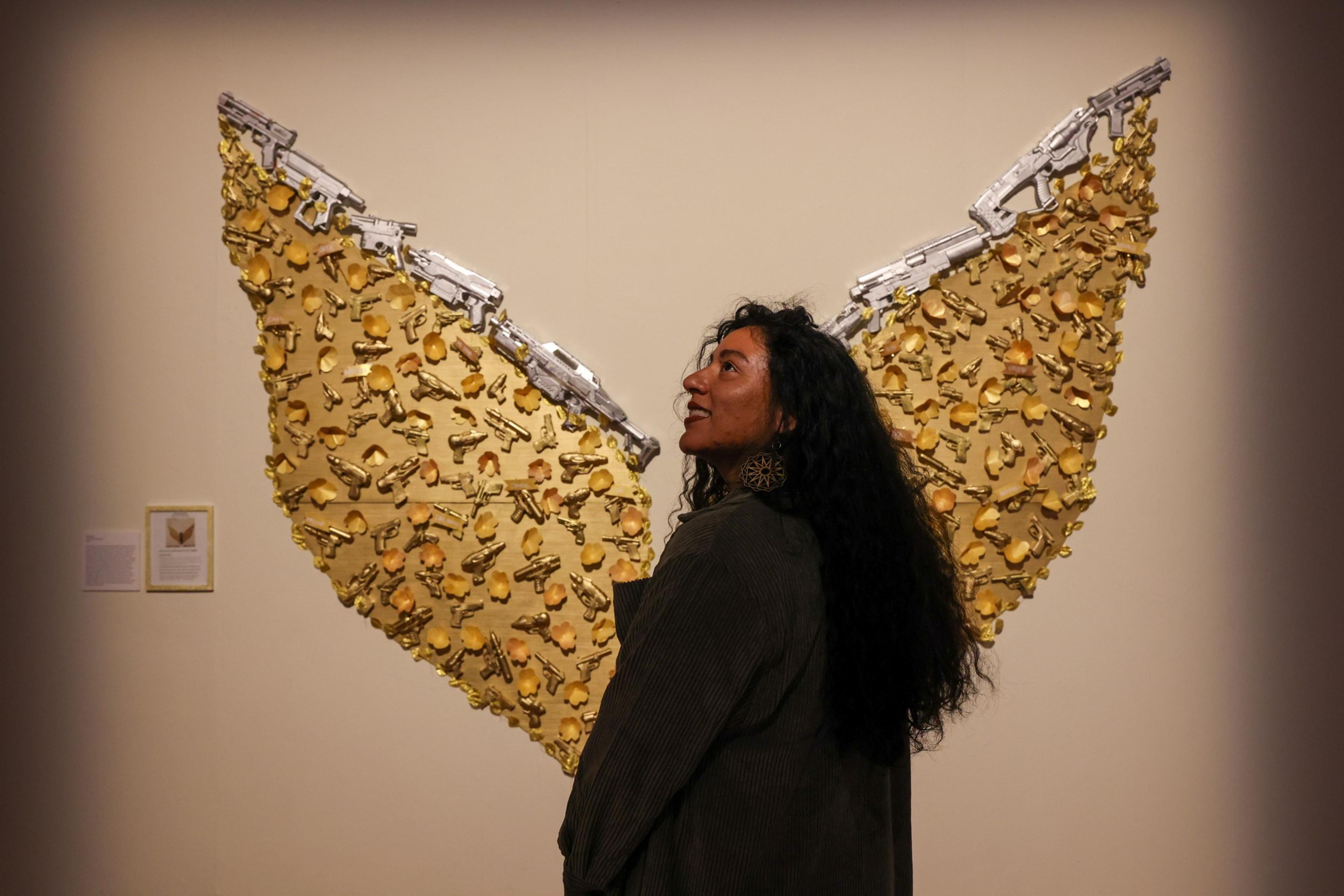 A woman with long dark hair stands in front of wall art shaped like wings, made of golden gun-shaped objects and silver toy guns at the top edges.