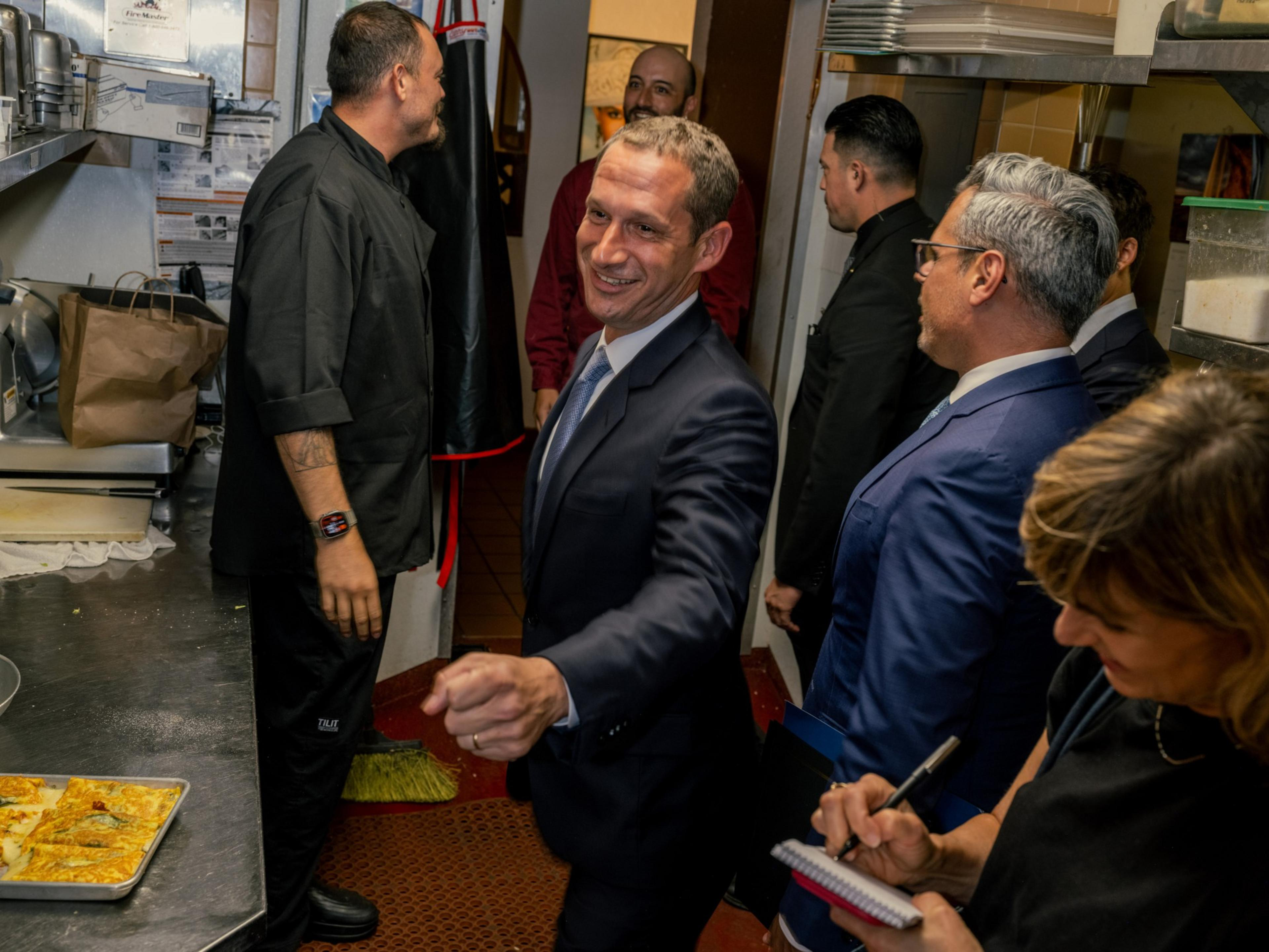 A man in a suit smiles and gestures in a busy kitchen while others, including a chef and a woman taking notes, stand around him.