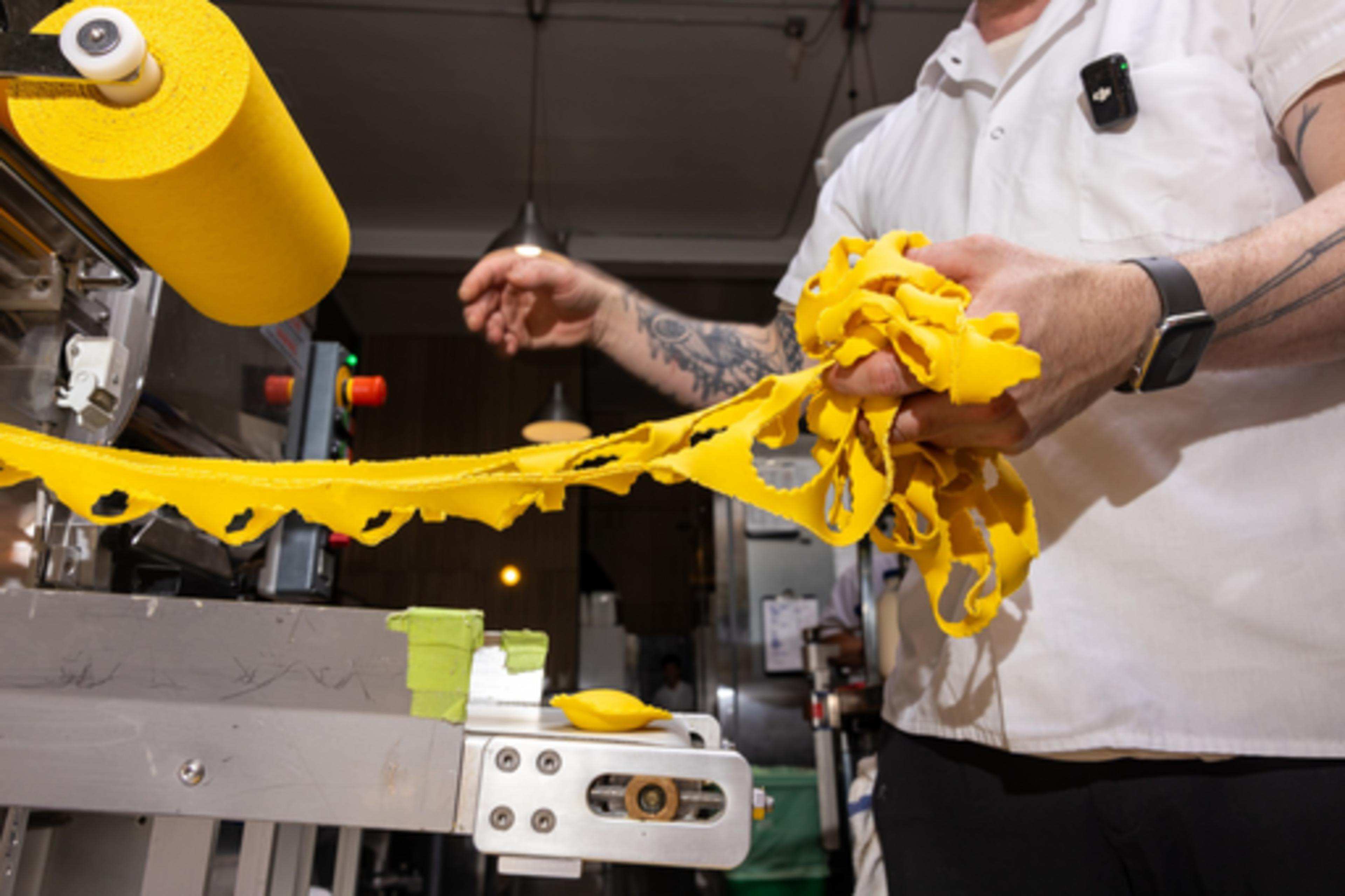 A person holds yellow pasta dough strands, shaped by a machine nearby, in an industrial kitchen. They wear a white shirt and have tattoos on their arms.