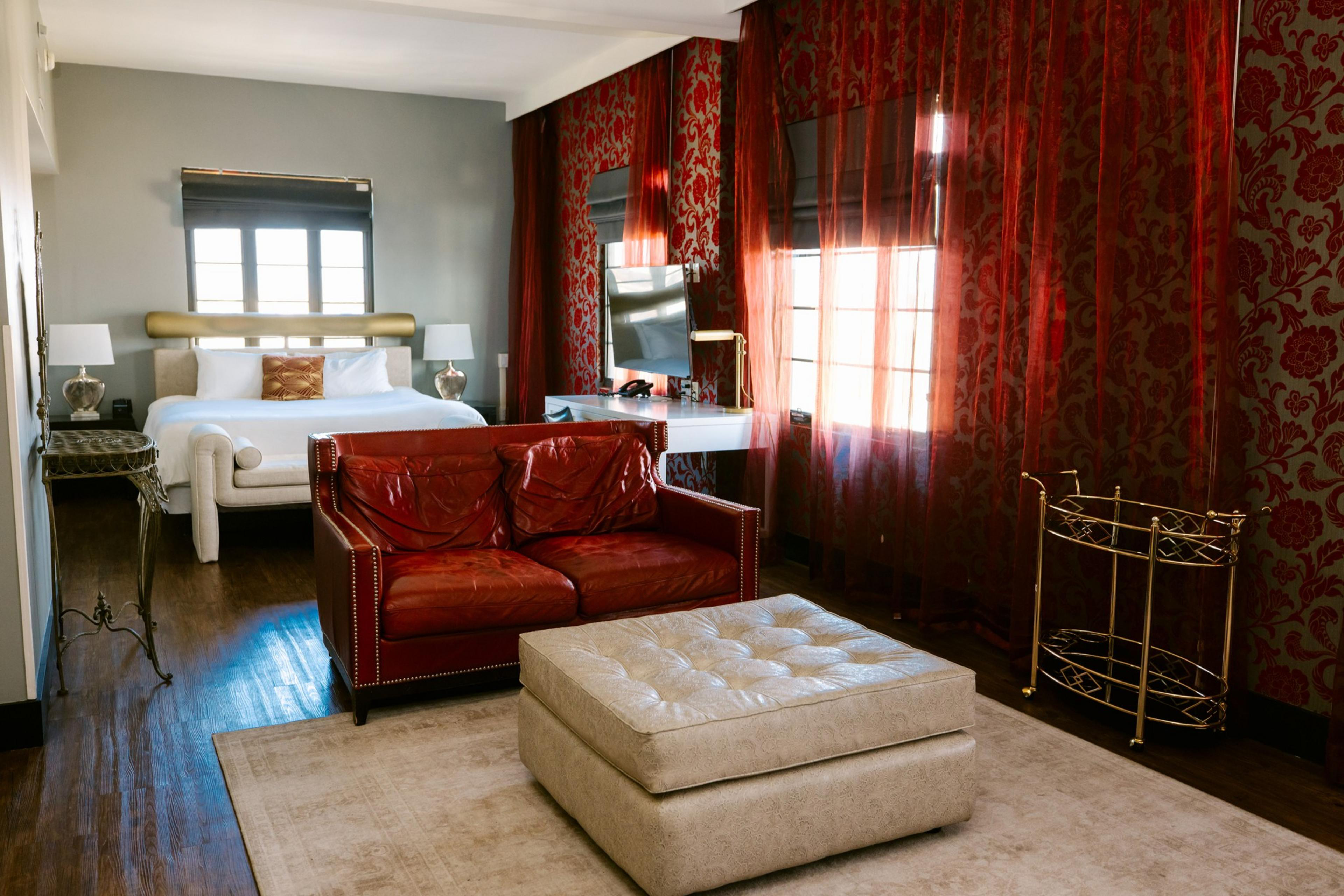 The image shows a stylish room with a large bed, red sofa, ottoman, and elegant decor. There's a TV, sheer curtains, and a cart against a textured red wall.