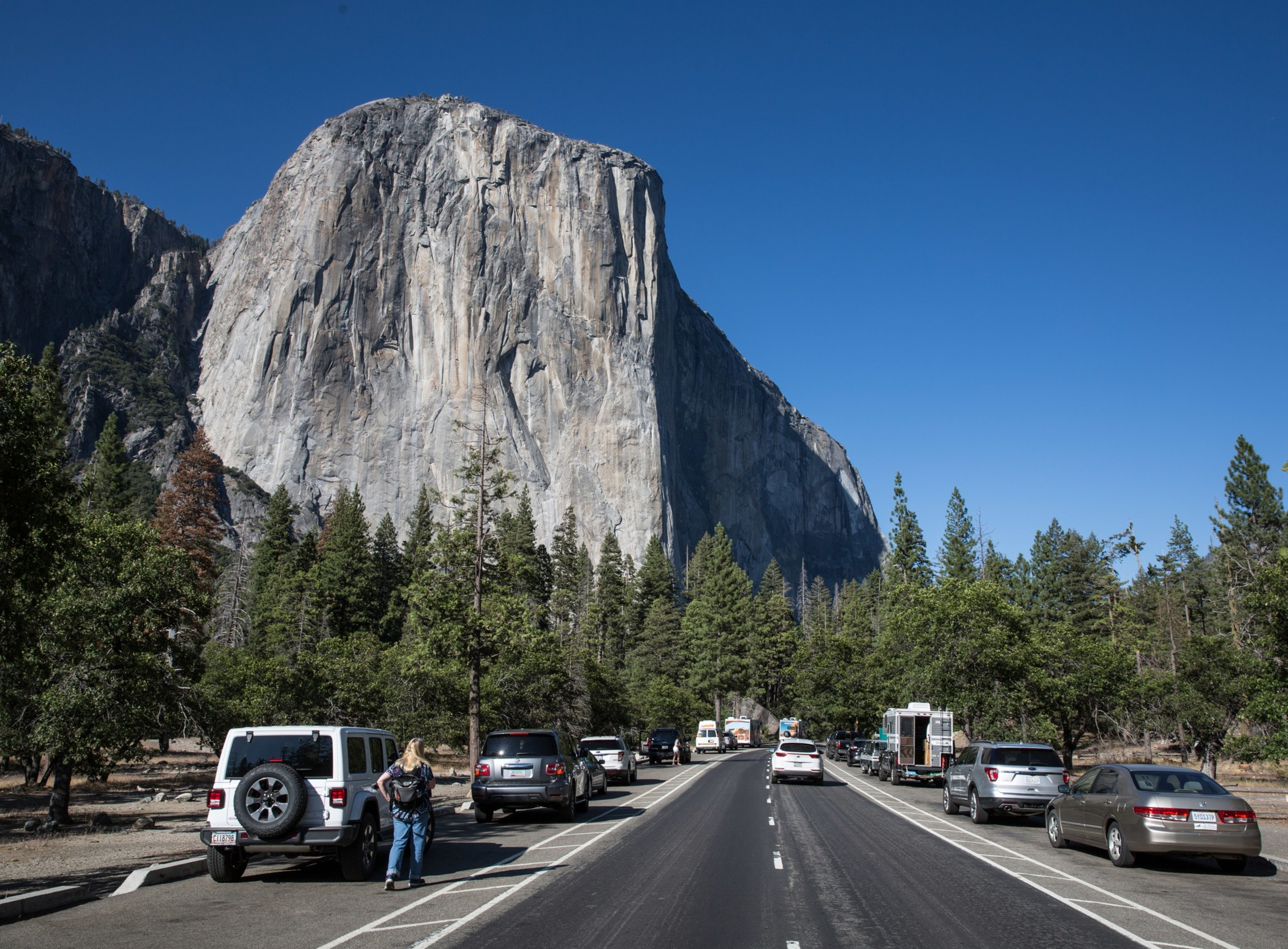 A towering rock formation stands under a clear blue sky, with a road lined by parked cars and surrounded by a forested area where a few people are visible.