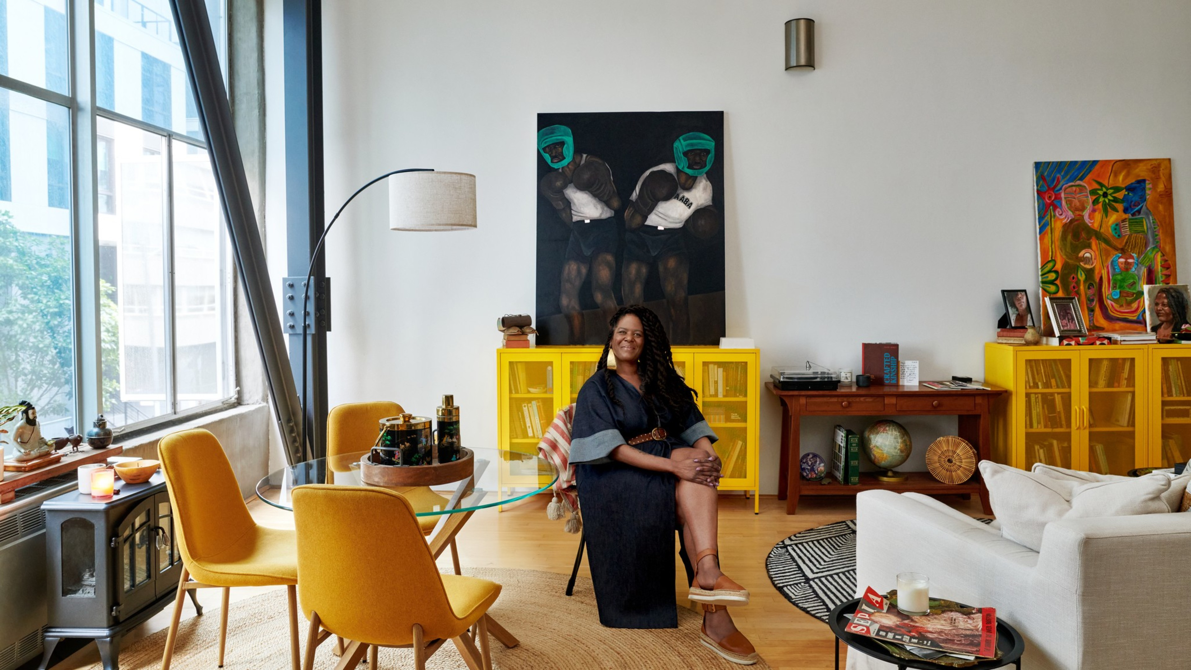 A person sits in a bright, art-filled living room with large windows, yellow chairs, a round glass table, and vibrant artwork, creating a cozy, stylish ambiance.