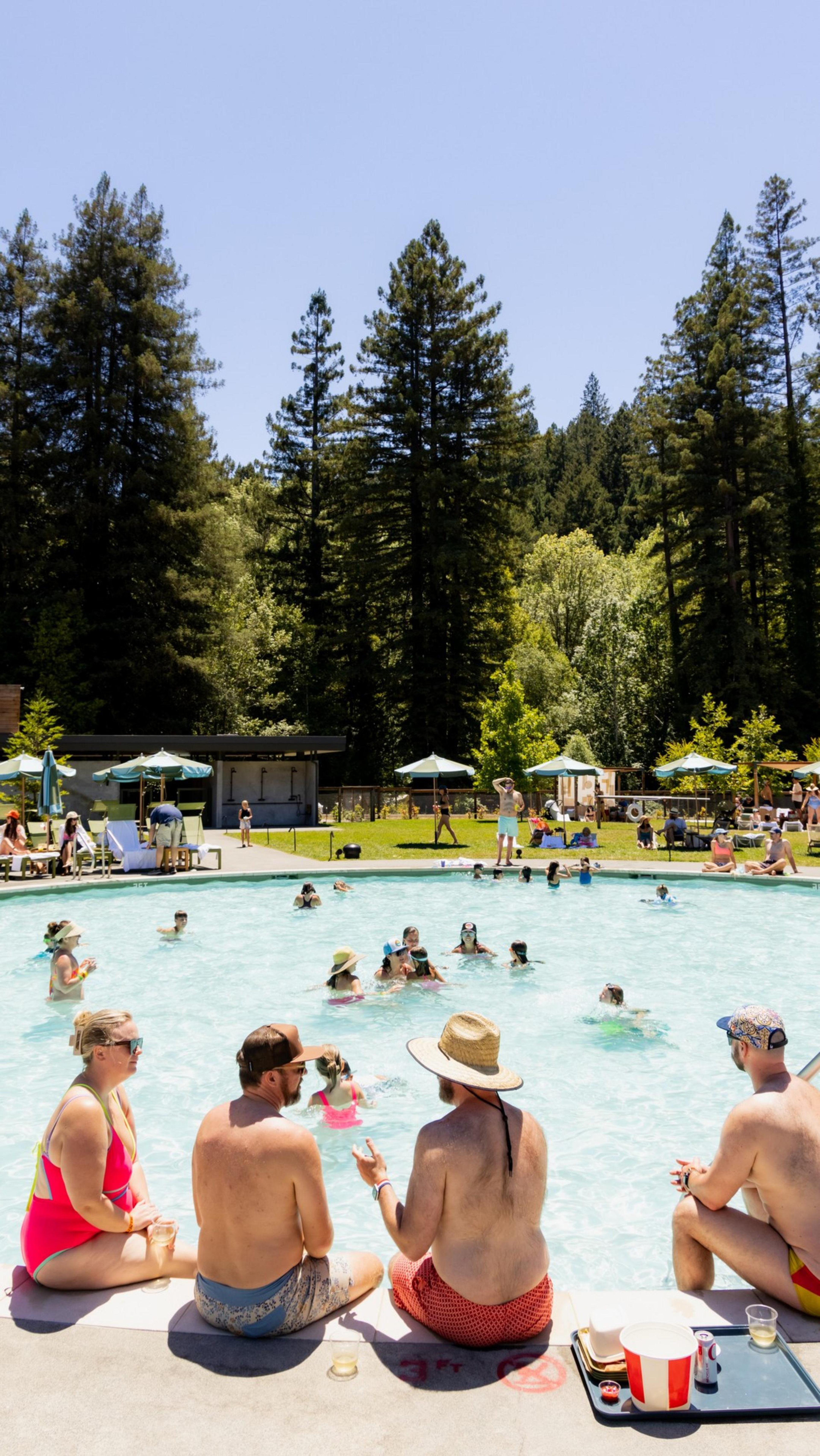 People are relaxing by a pool surrounded by tall trees. Some sit by the edge with drinks, while others swim. Green umbrellas and a building are in the background.