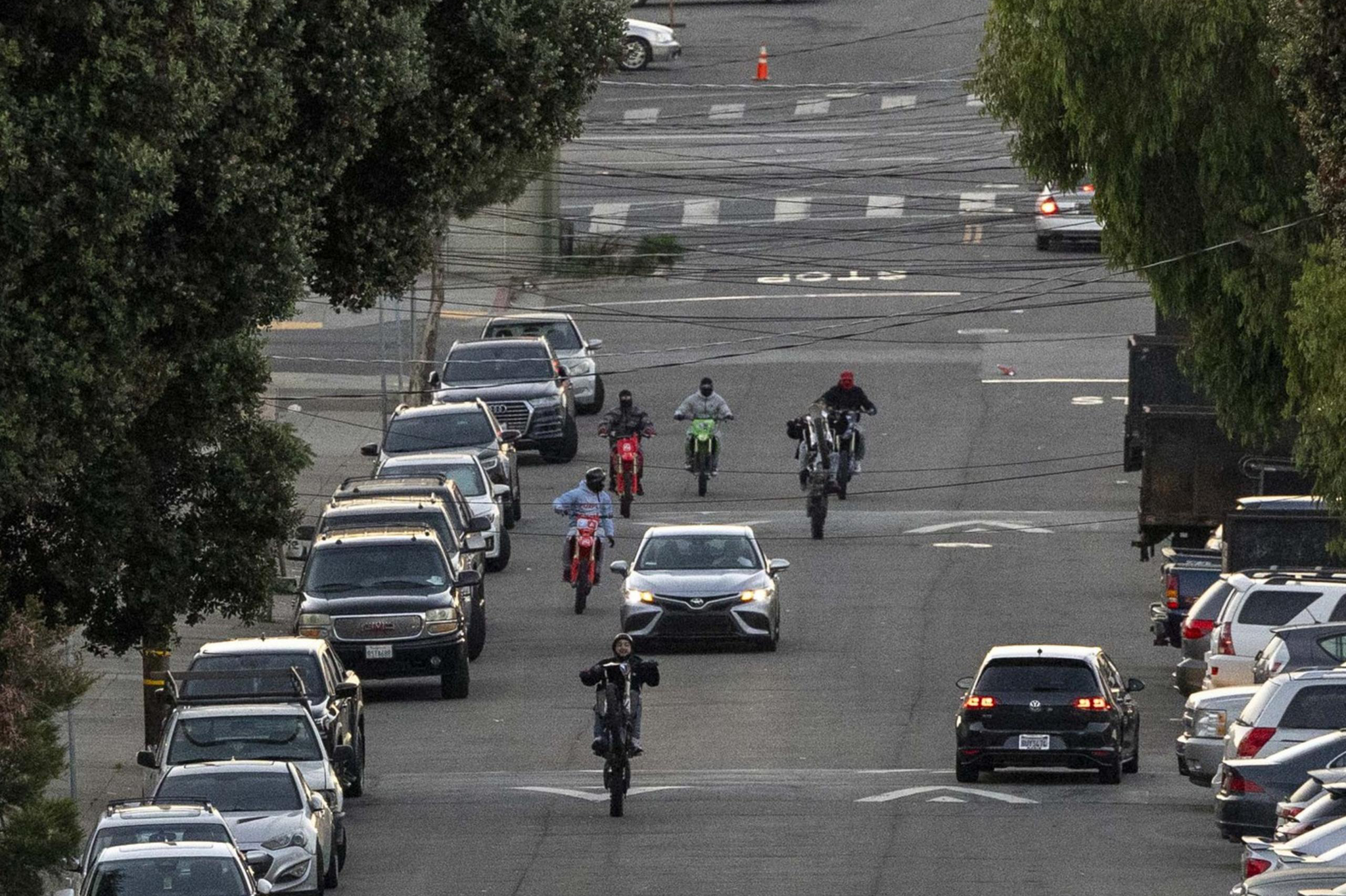 A group of motorcyclists rides up a sloping urban street, some performing wheelies. Cars are parked on both sides, and trees line the sidewalks.