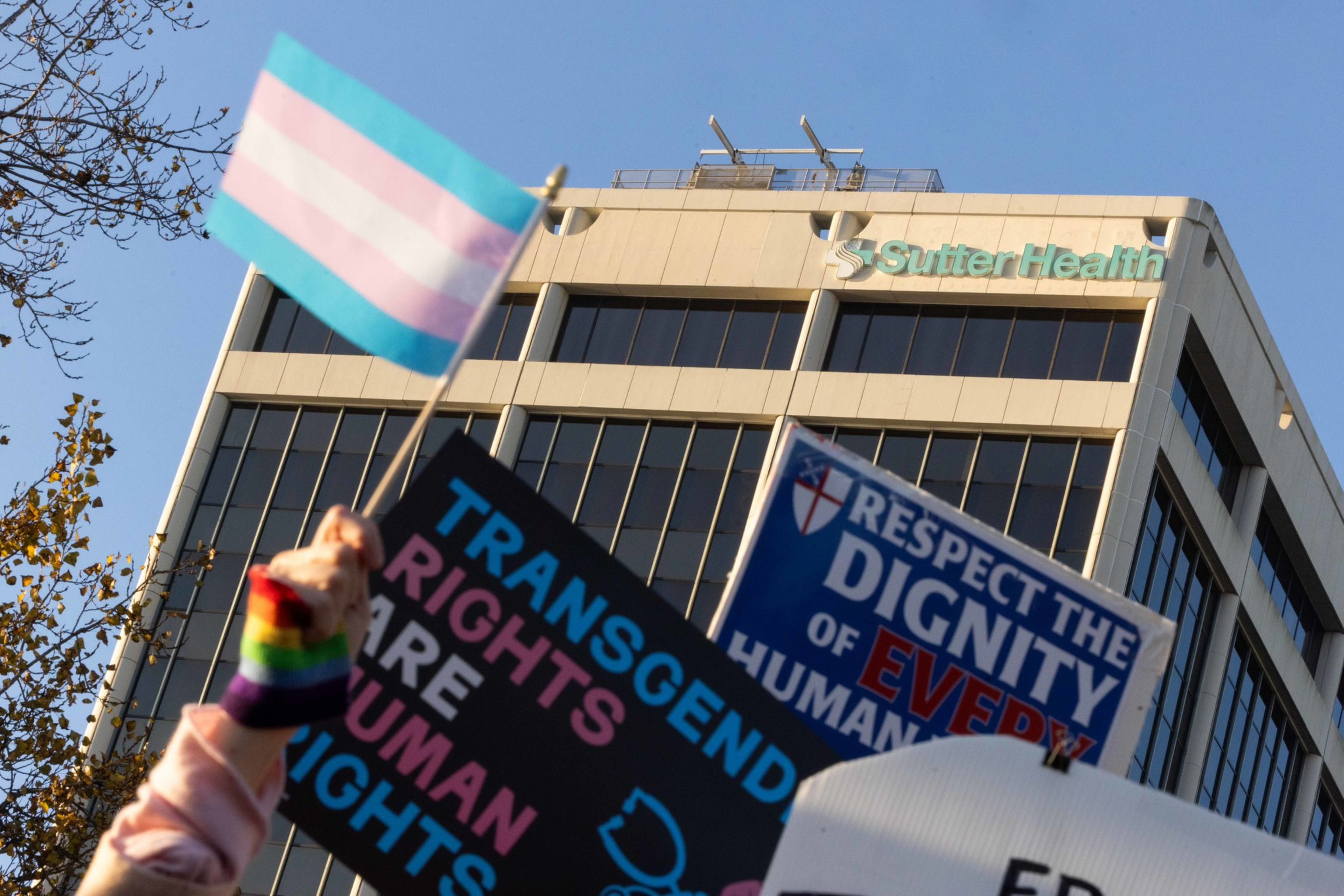 A person holds a transgender pride flag and rainbow wristbands along with signs reading &quot;Transgender rights are human rights&quot; and &quot;Respect the dignity of every human&quot; near a Sutter Health building.