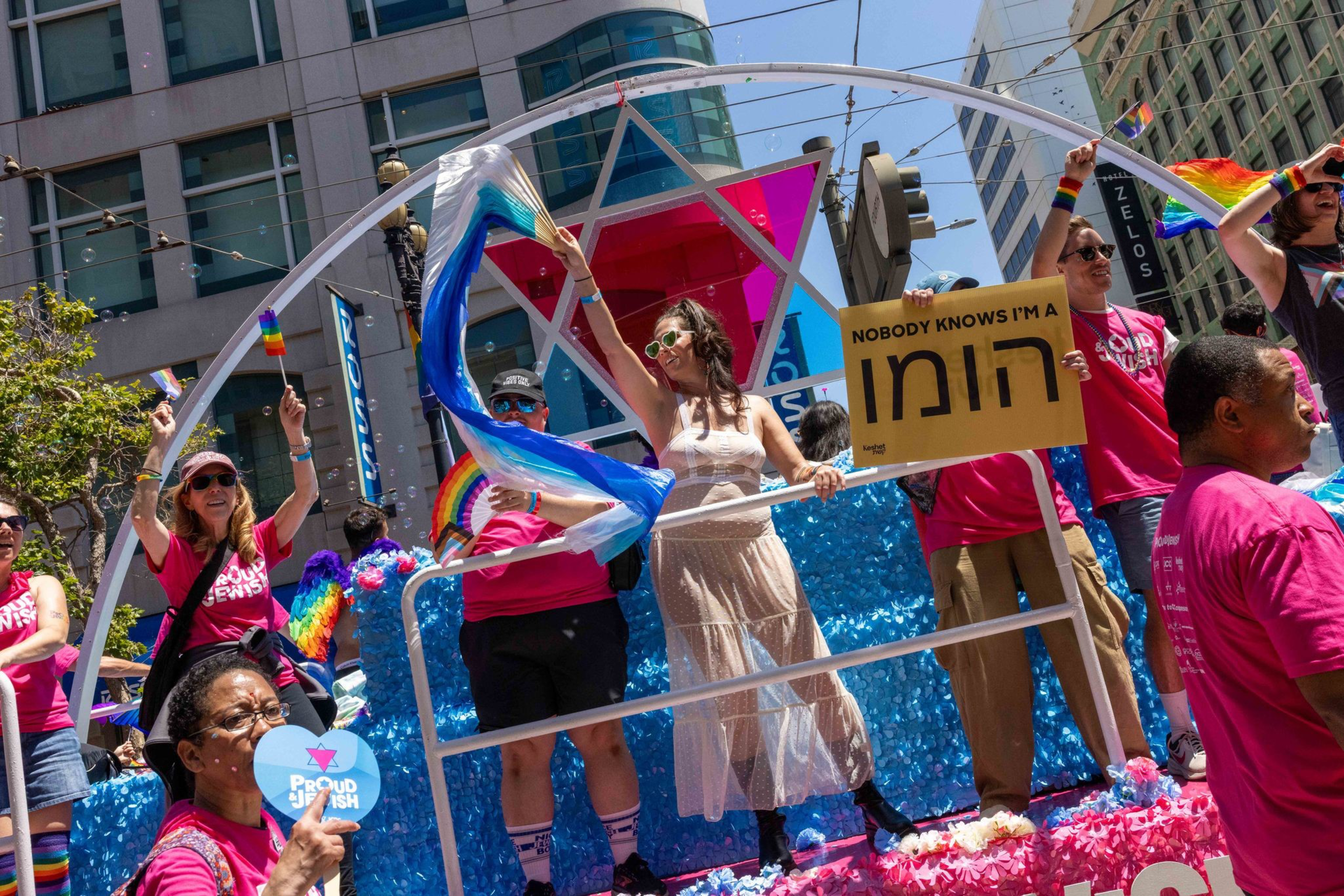People on a colorful parade float are cheerfully waving pride flags, with &quot;Proud Jewish&quot; shirts and a sign saying &quot;Nobody knows I'm a [Hebrew word].&quot;