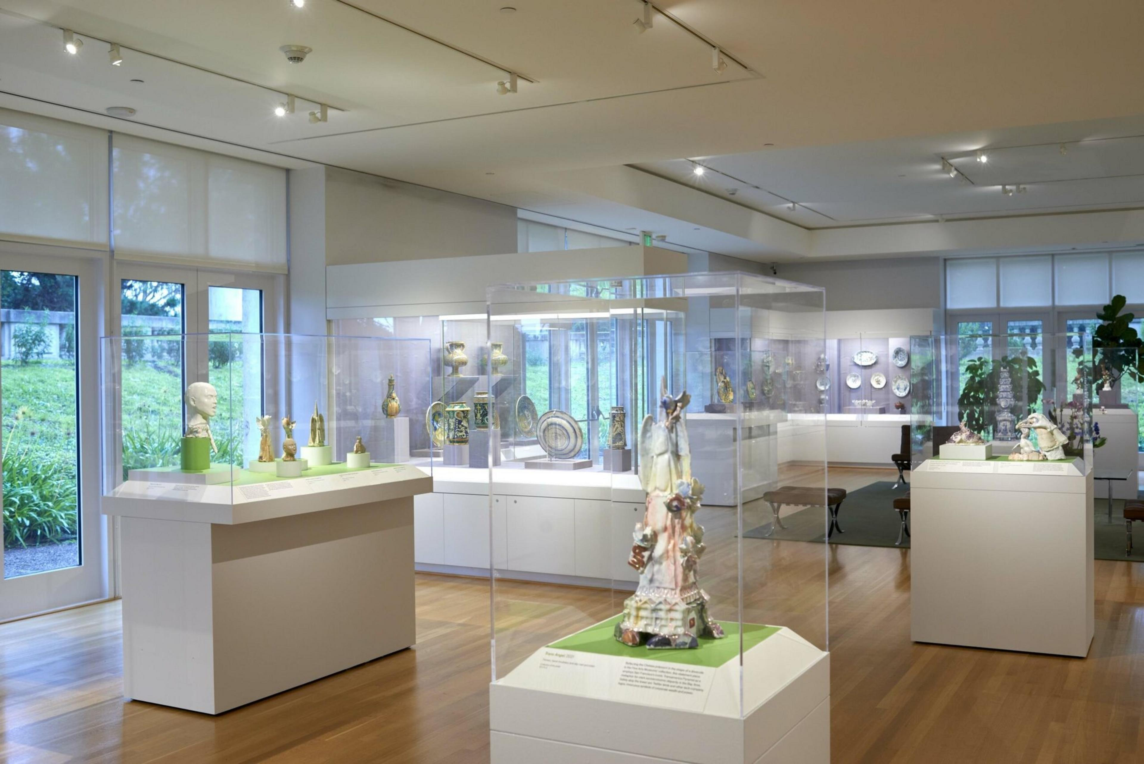 A bright, modern museum gallery displays various ceramics and sculptures in glass cases on white pedestals, with large windows and wooden floors.