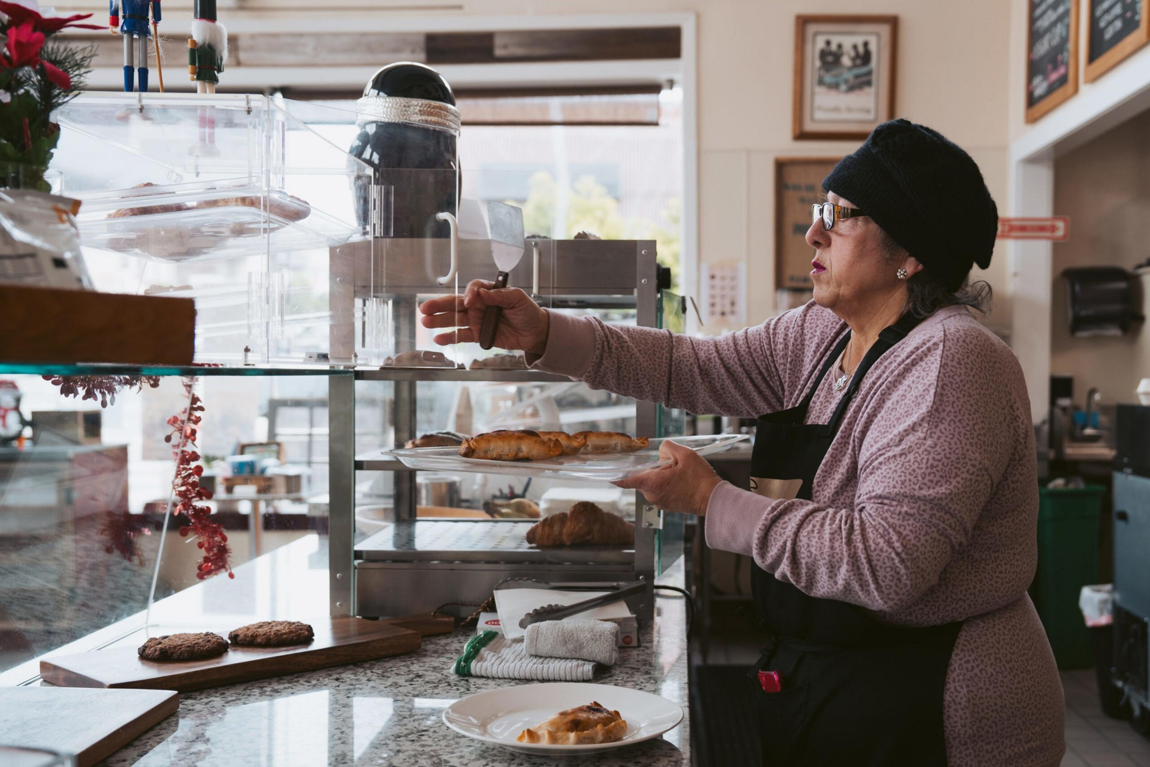 A woman in a black beanie and glasses uses a spatula to serve pastries from a glass display case onto a plate in a bakery.
