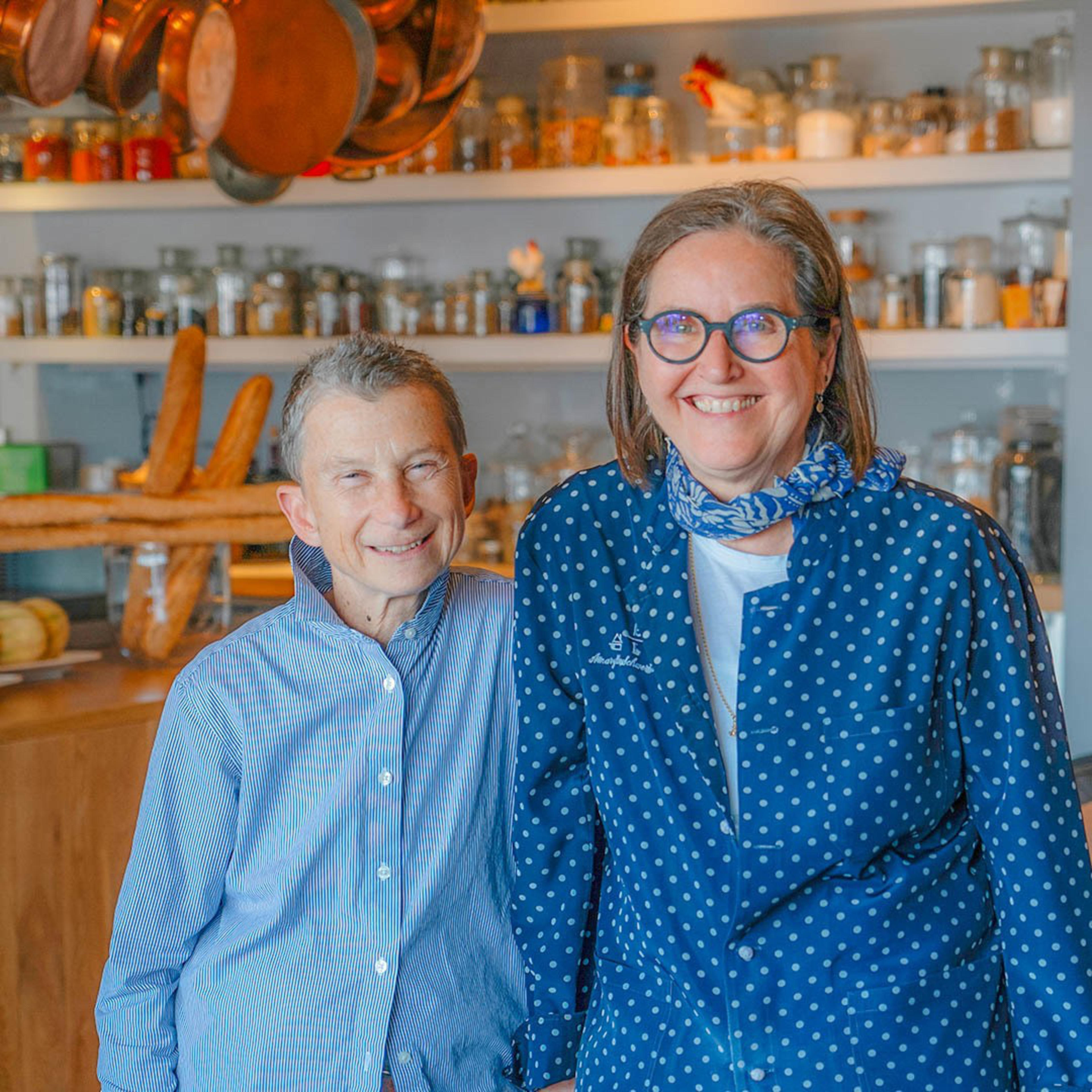 Two smiling people stand close together in a kitchen filled with jars, bread, and hanging pots, wearing blue-patterned shirts and glasses.