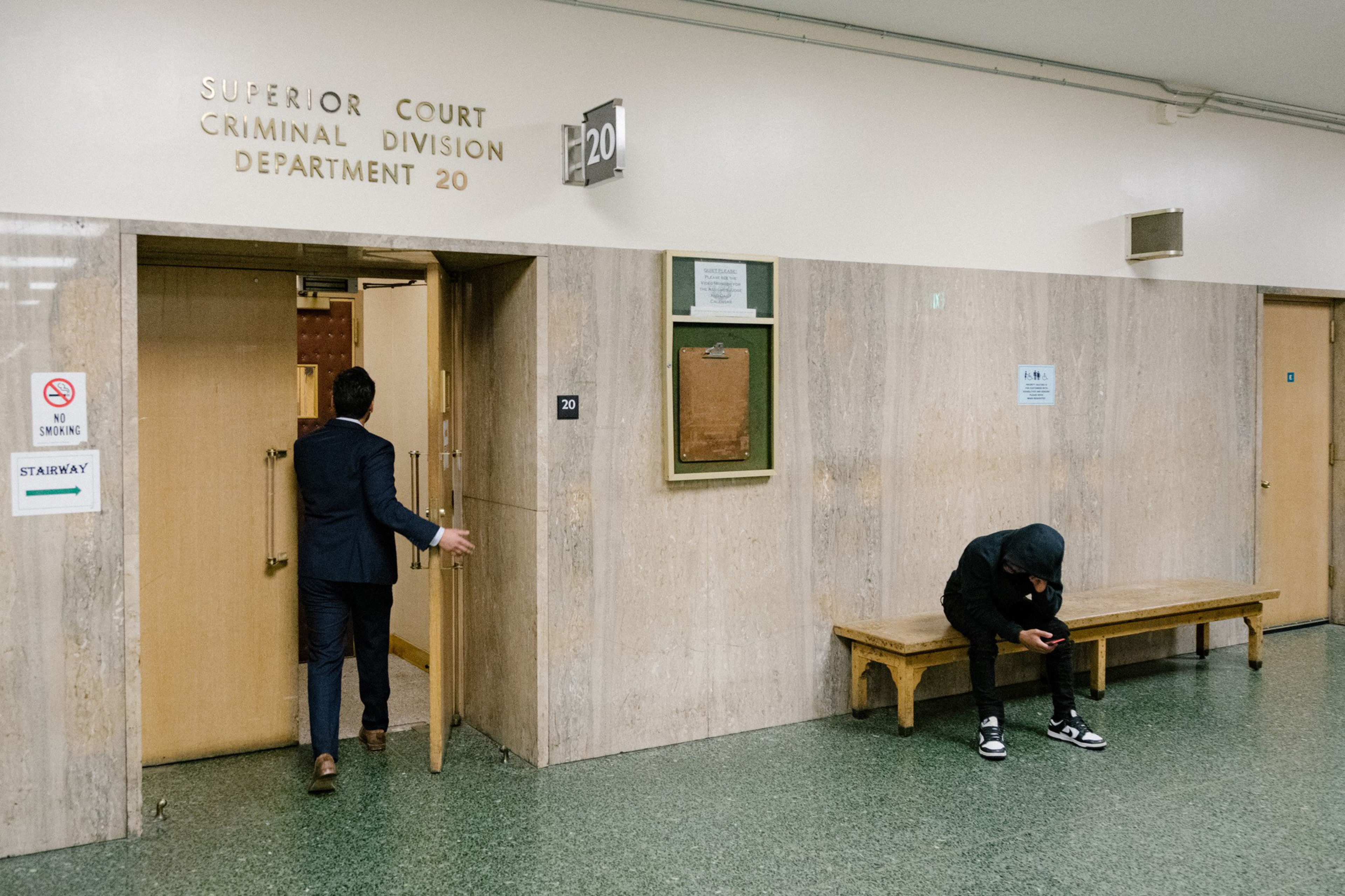 A man in a suit enters a courtroom door marked "Superior Court Criminal Division Department 20," while a hooded person sits on a bench looking at a phone.