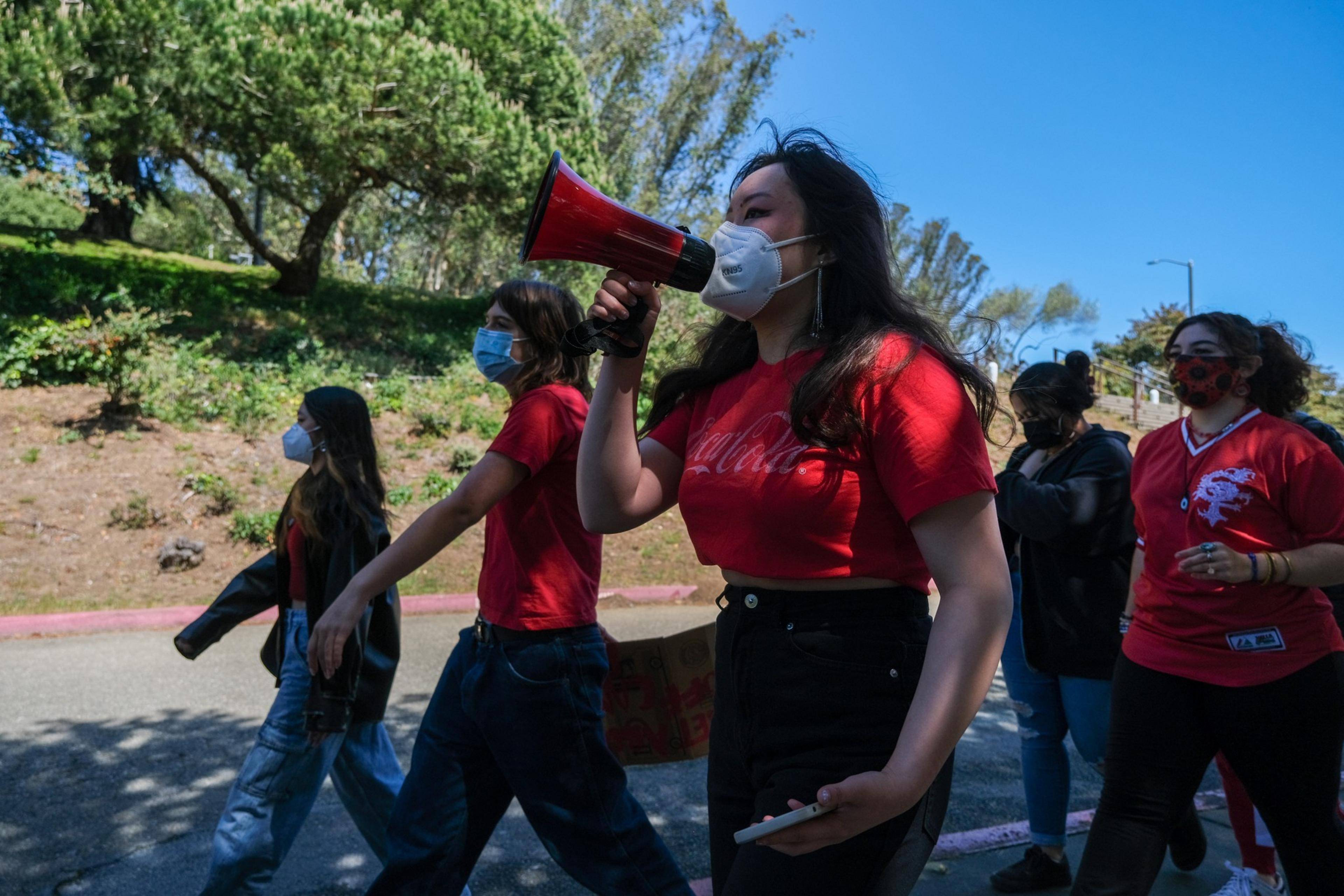 A group of people wearing masks walks outdoors, with one woman in a red shirt speaking into a red megaphone.
