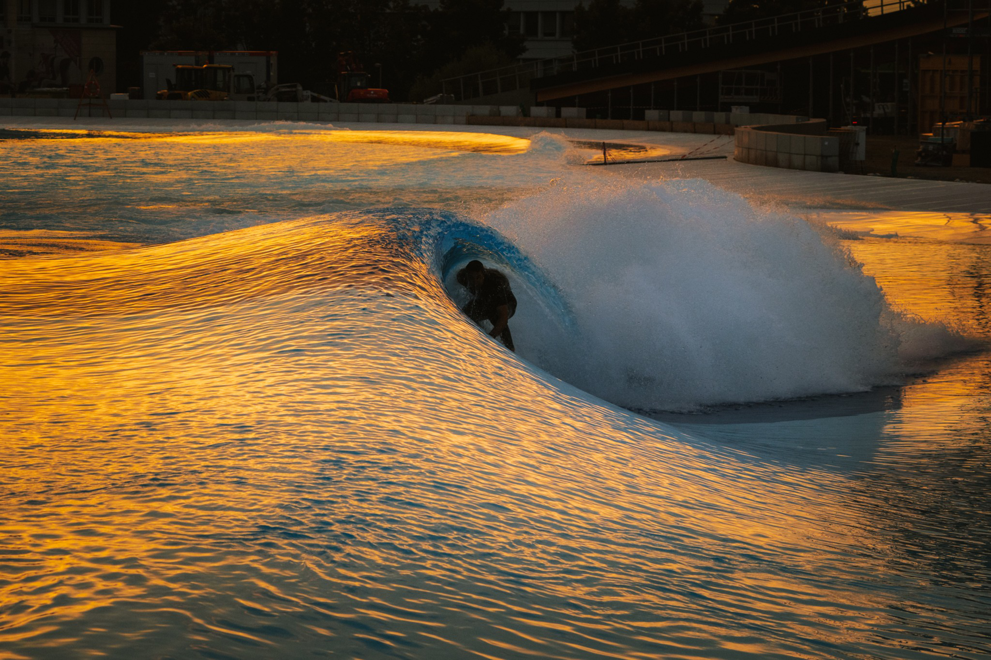 A surfer rides a glowing golden wave, illuminated by the warm light of a setting sun. The wave forms a tube around them as water splashes nearby.