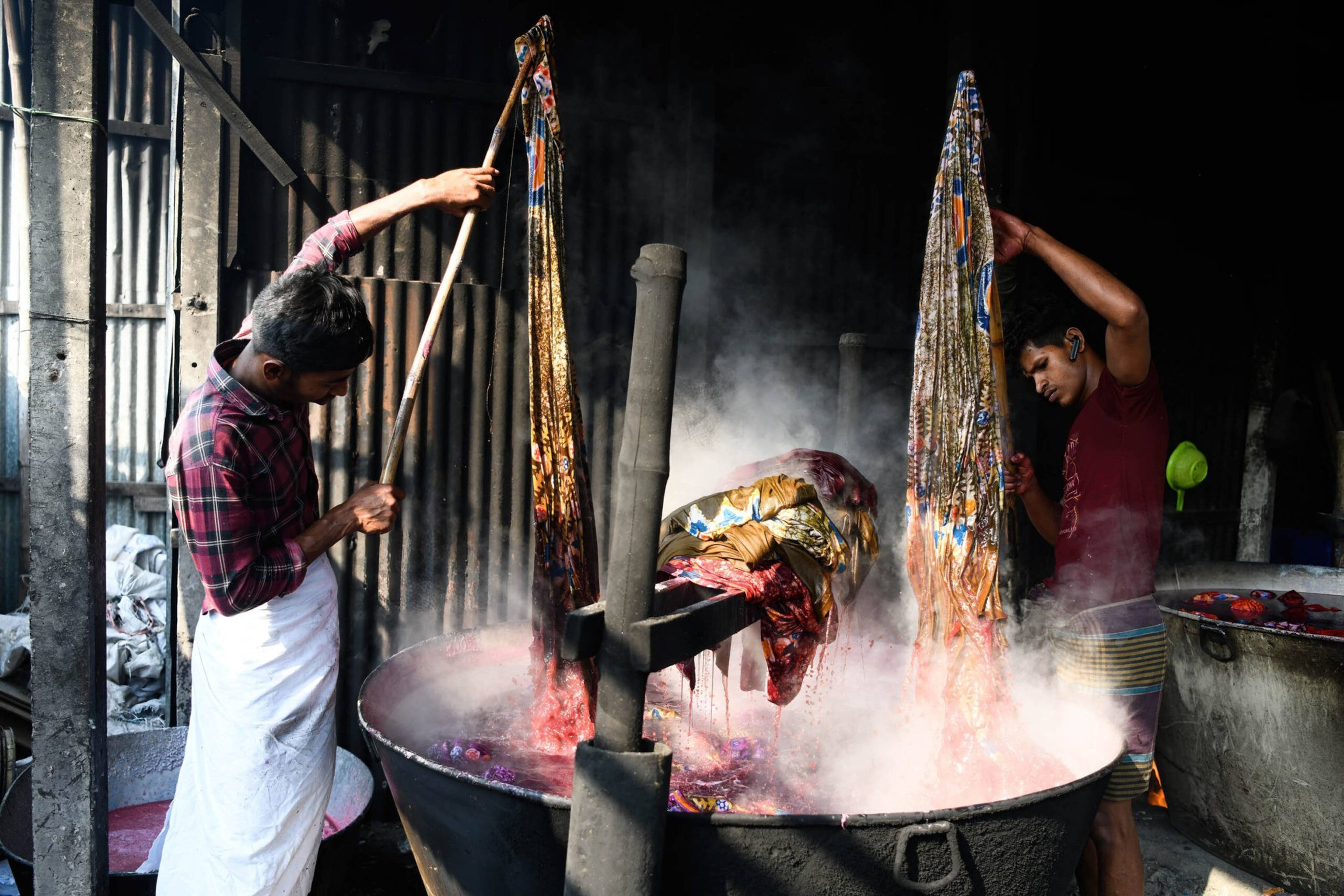Two men are dyeing fabric in large steaming vats, surrounded by a rustic workshop setting.